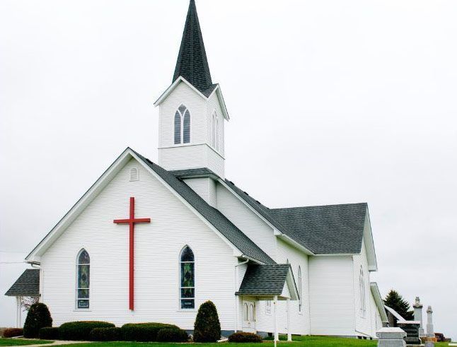 White church with red cross, steeple, and dark roof under a cloudy sky.