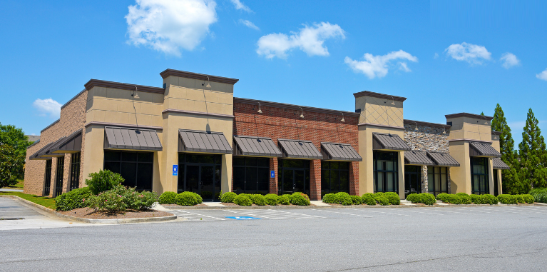 Building with tan and brick facade, awnings, and large windows, against a blue sky.