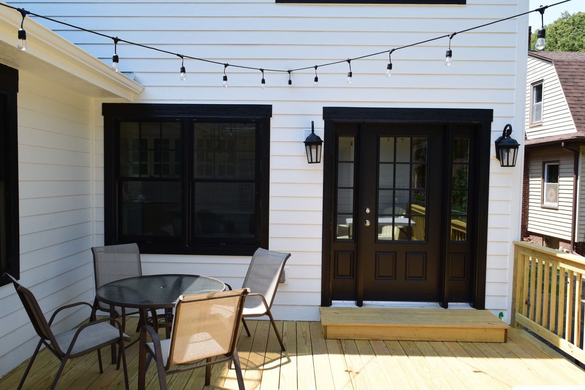 White house exterior with black trim, a deck, and string lights over a seating area with a table and chairs.