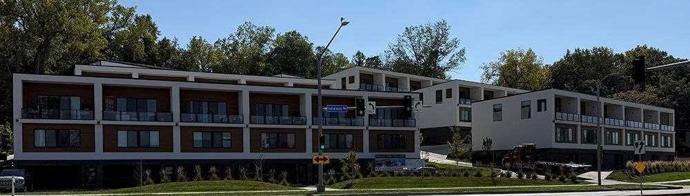 Modern apartment building with balconies, brown and white facade, and lush green trees in the background.