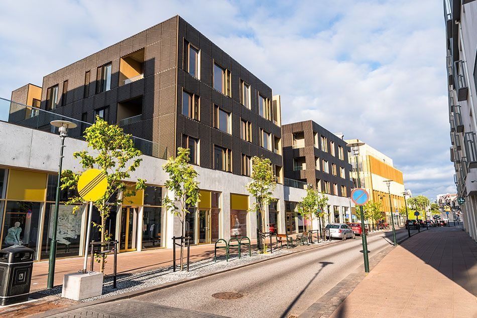 Street view with modern buildings, shops with yellow accents, trees, and parked cars on a sunny day.