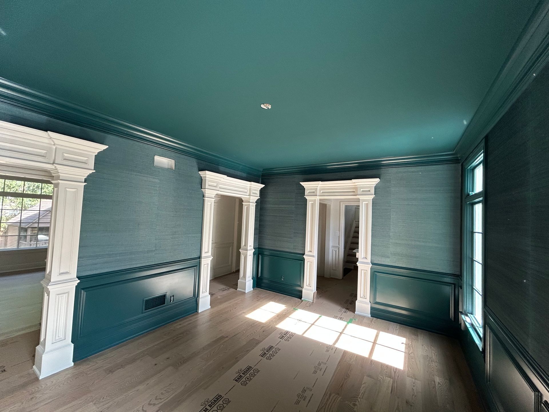 Empty room with teal walls, crown molding, and wooden floors. Sunlight streams through the windows.
