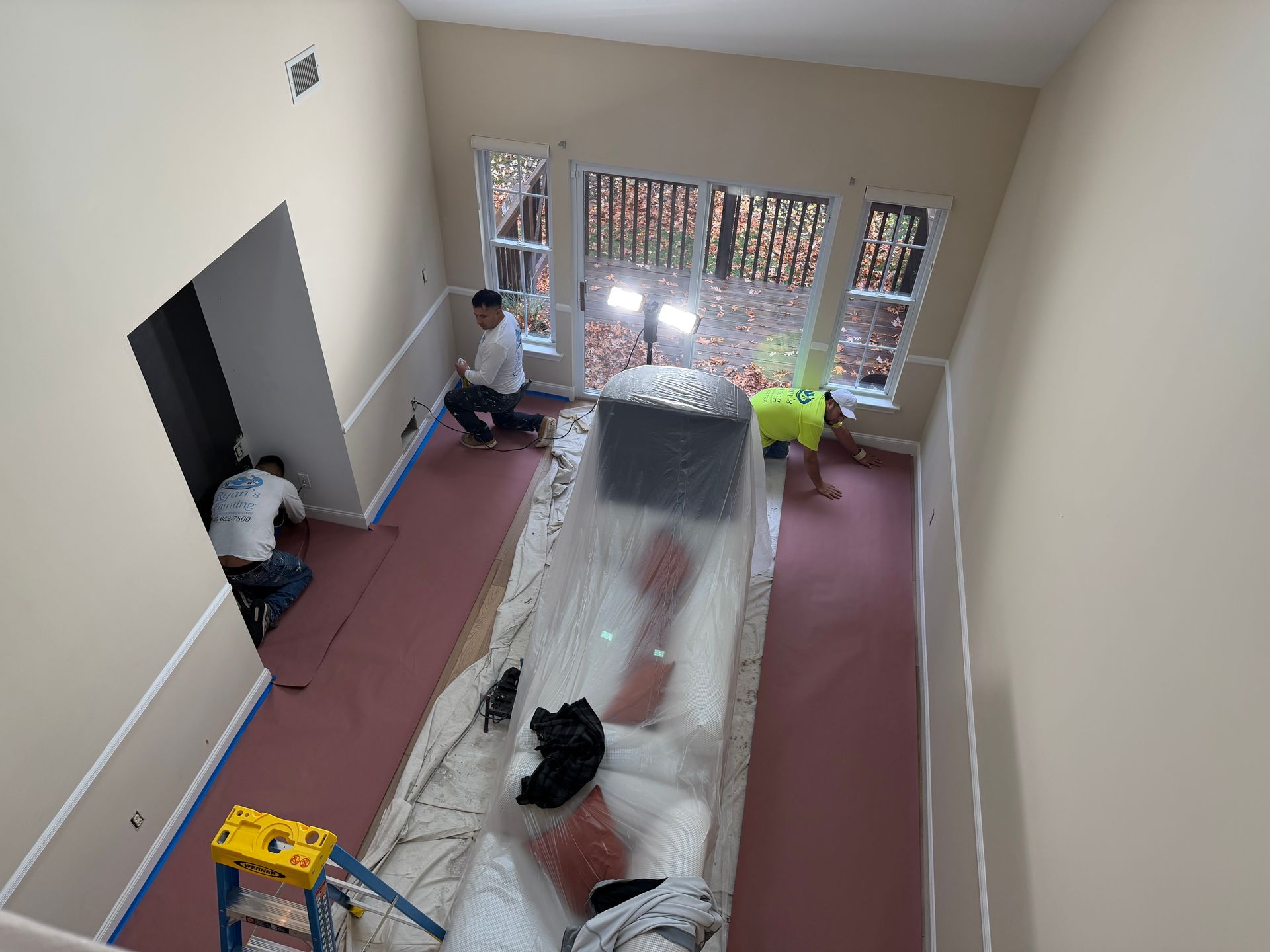Workers install flooring in a room with tall windows. Protective coverings and tools are visible.