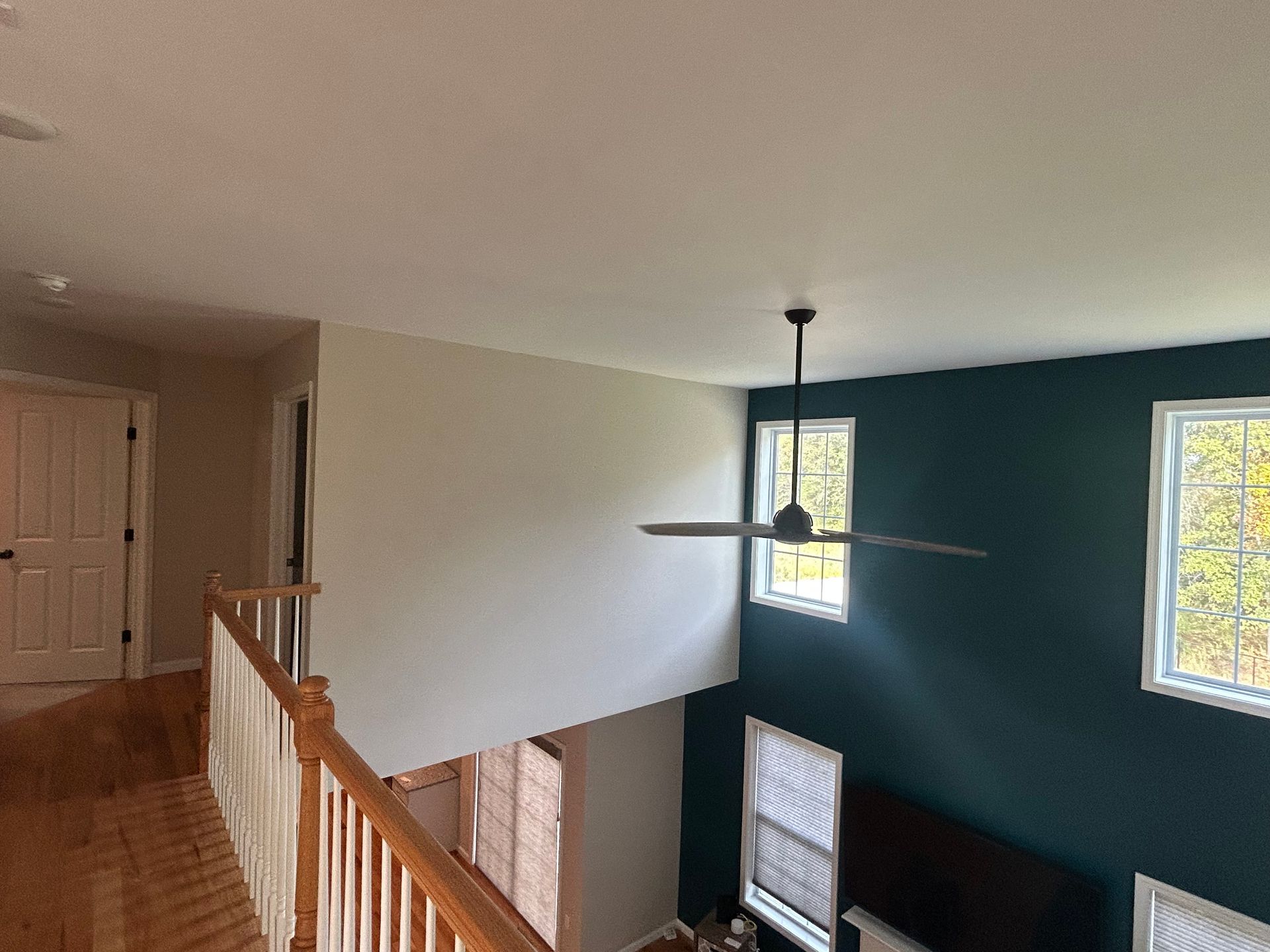 Loft interior: beige and teal walls, wooden railing, ceiling fan, and windows overlooking greenery.