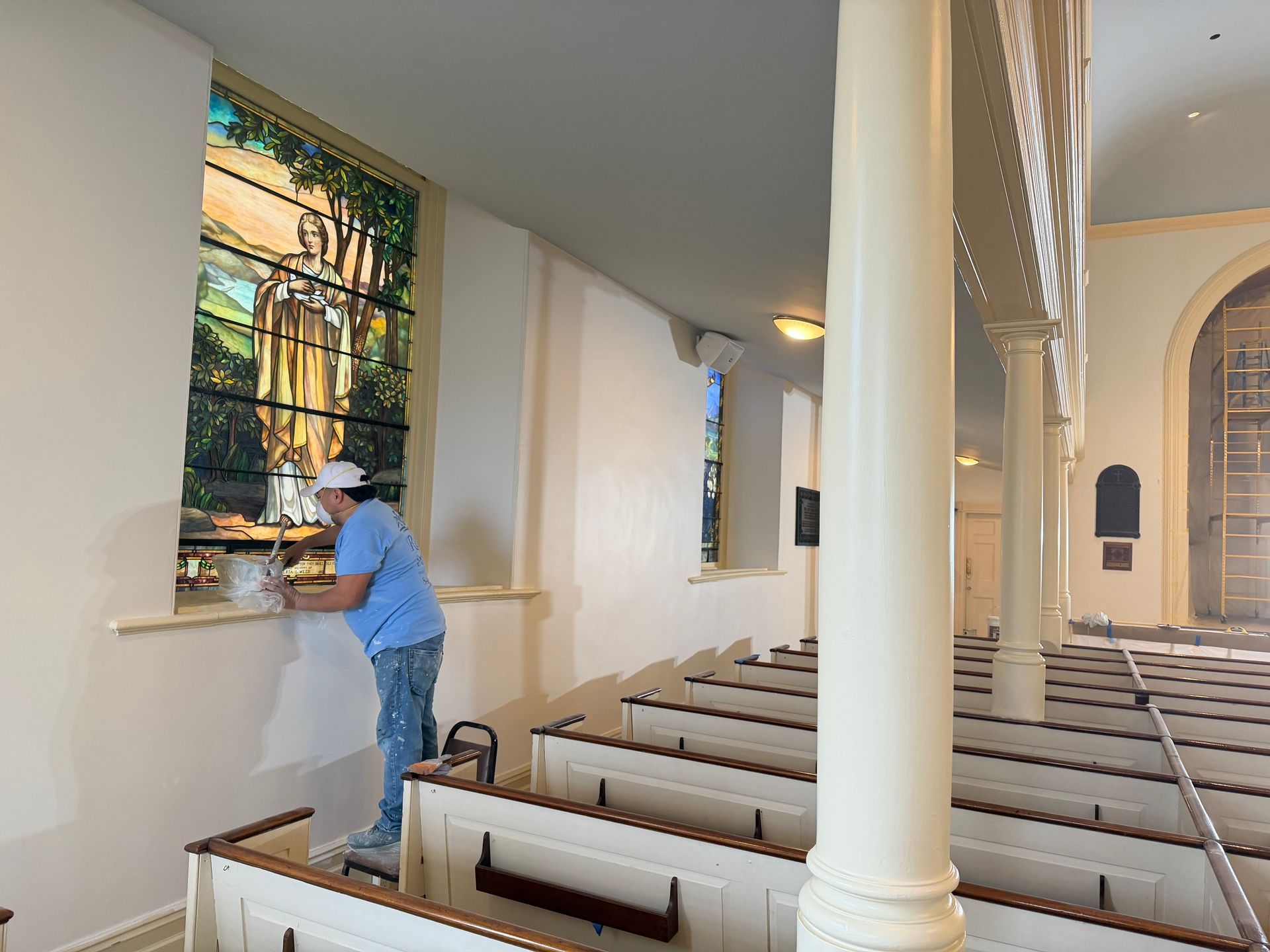 Person painting a wall in a church with pews and a stained glass window.