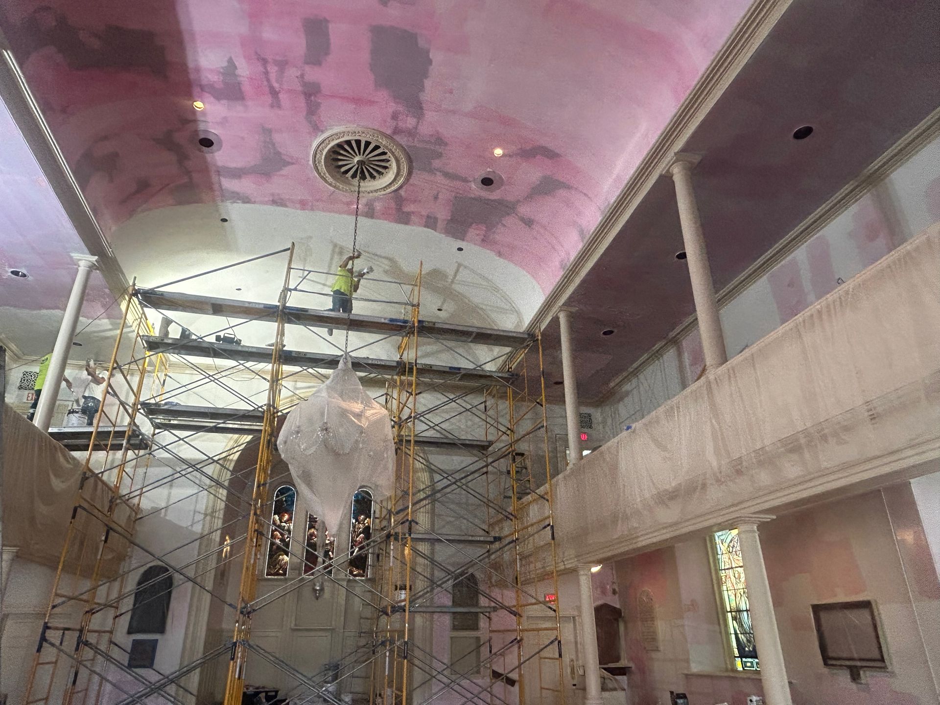 Interior construction of a church with scaffolding, workers on the ceiling, and a chandelier covered in plastic.