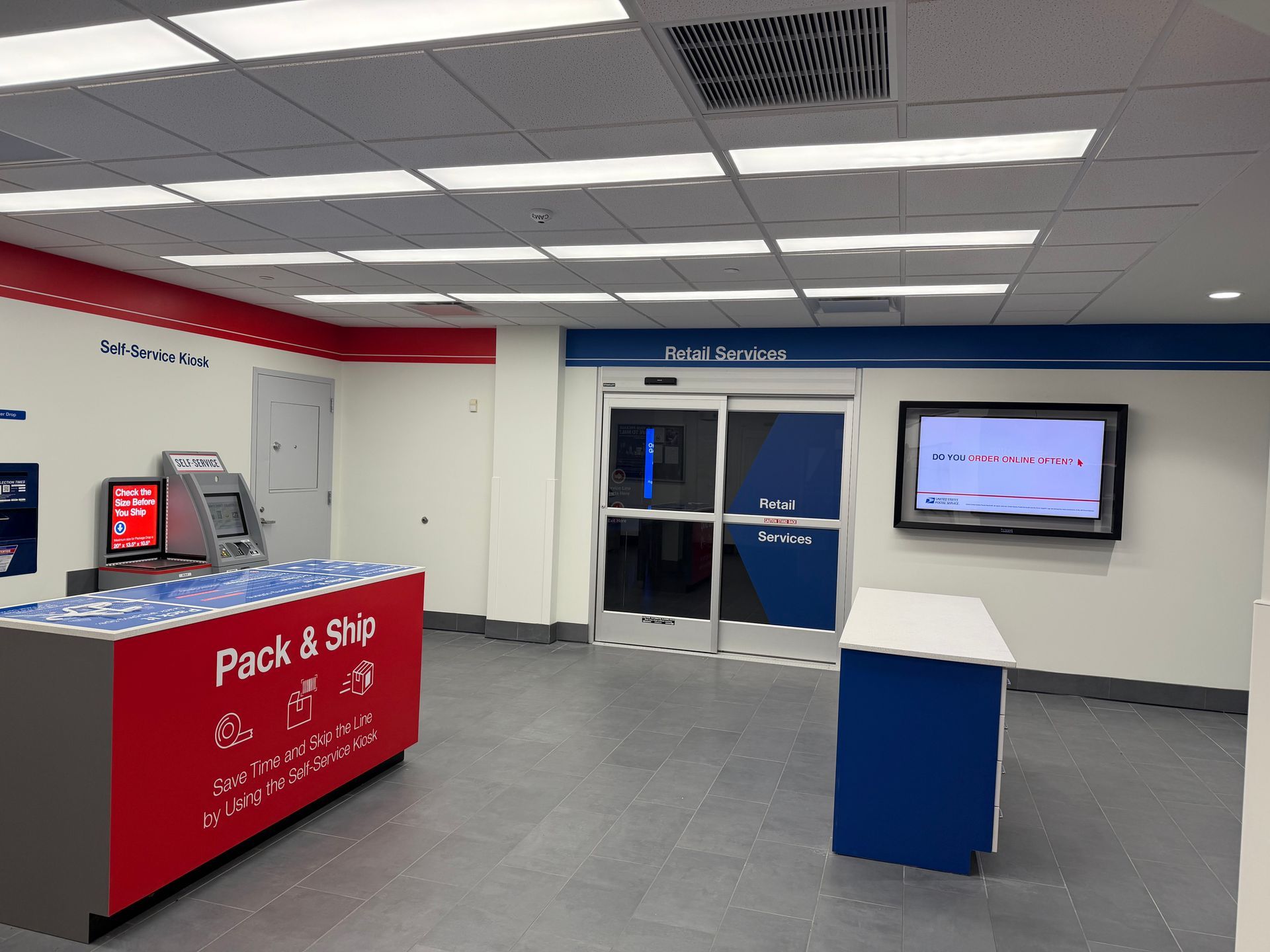 Interior of a USPS office: Pack & Ship station, ATM, entrance, and TV on wall; red, white, blue color scheme.
