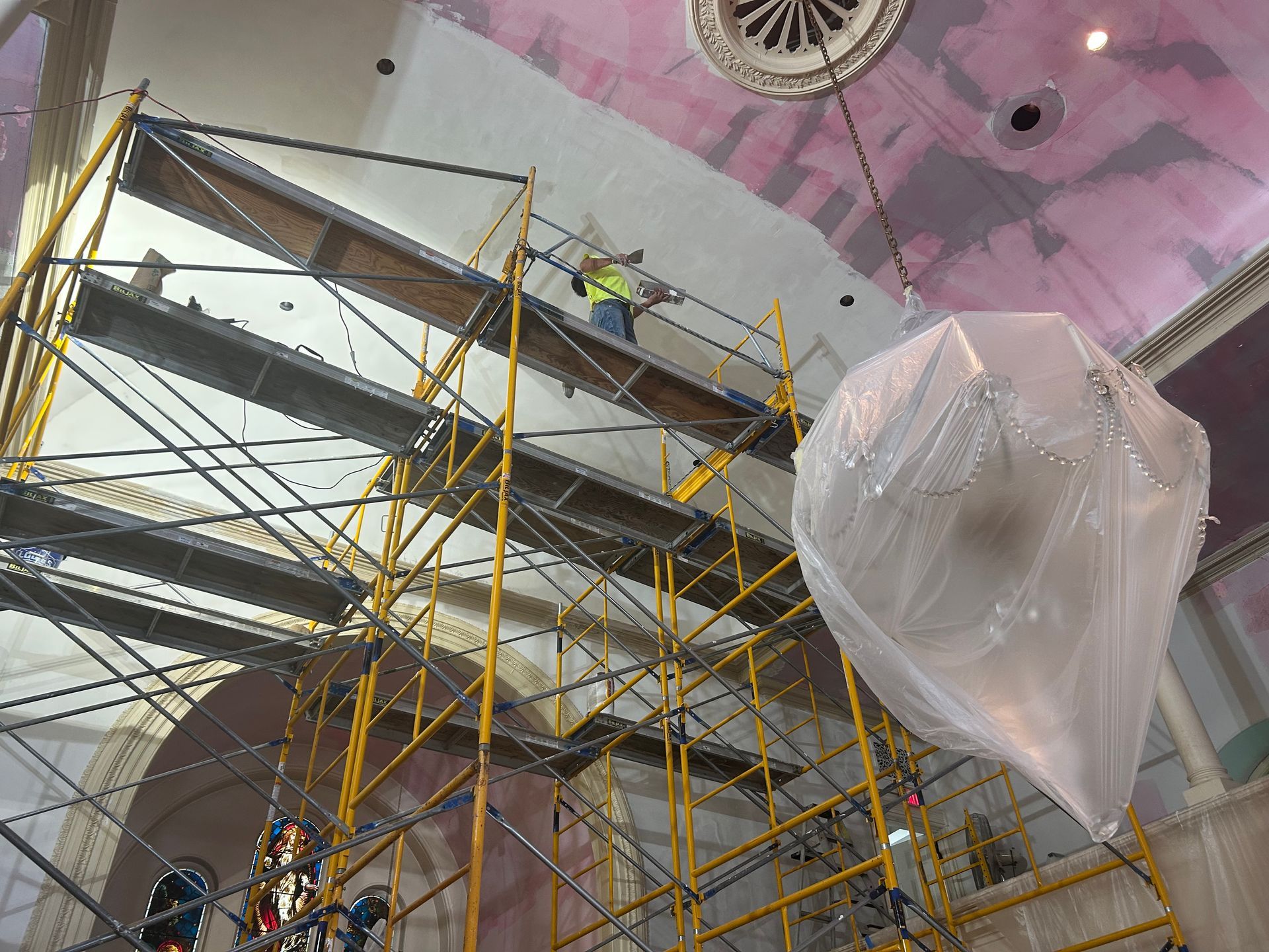 A worker on yellow scaffolding repairs a high ceiling in a light-colored interior; a large plastic covering hangs.