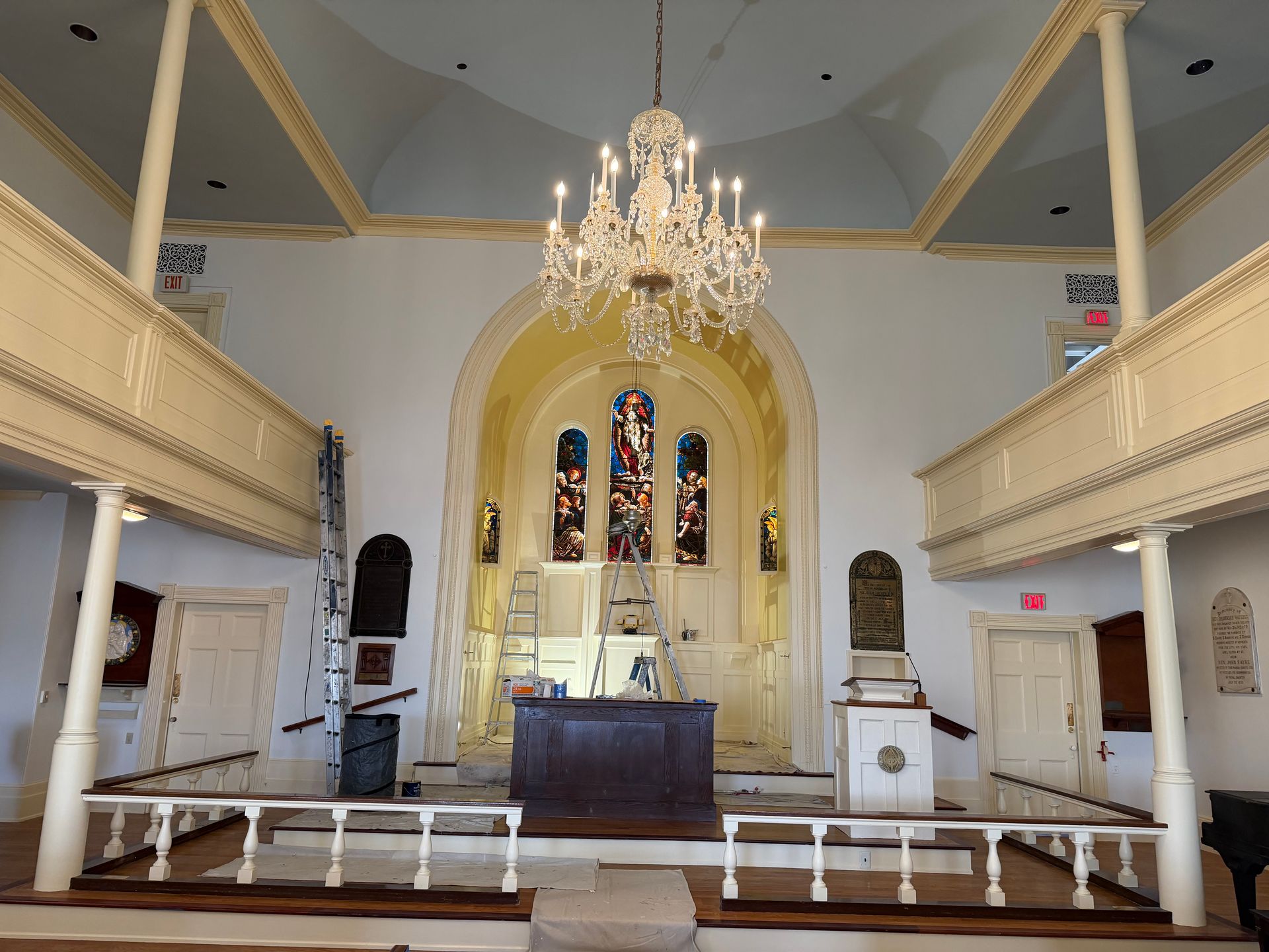 Interior of a church: stained glass, ornate chandelier, balcony on each side, wooden pulpit, and light walls.