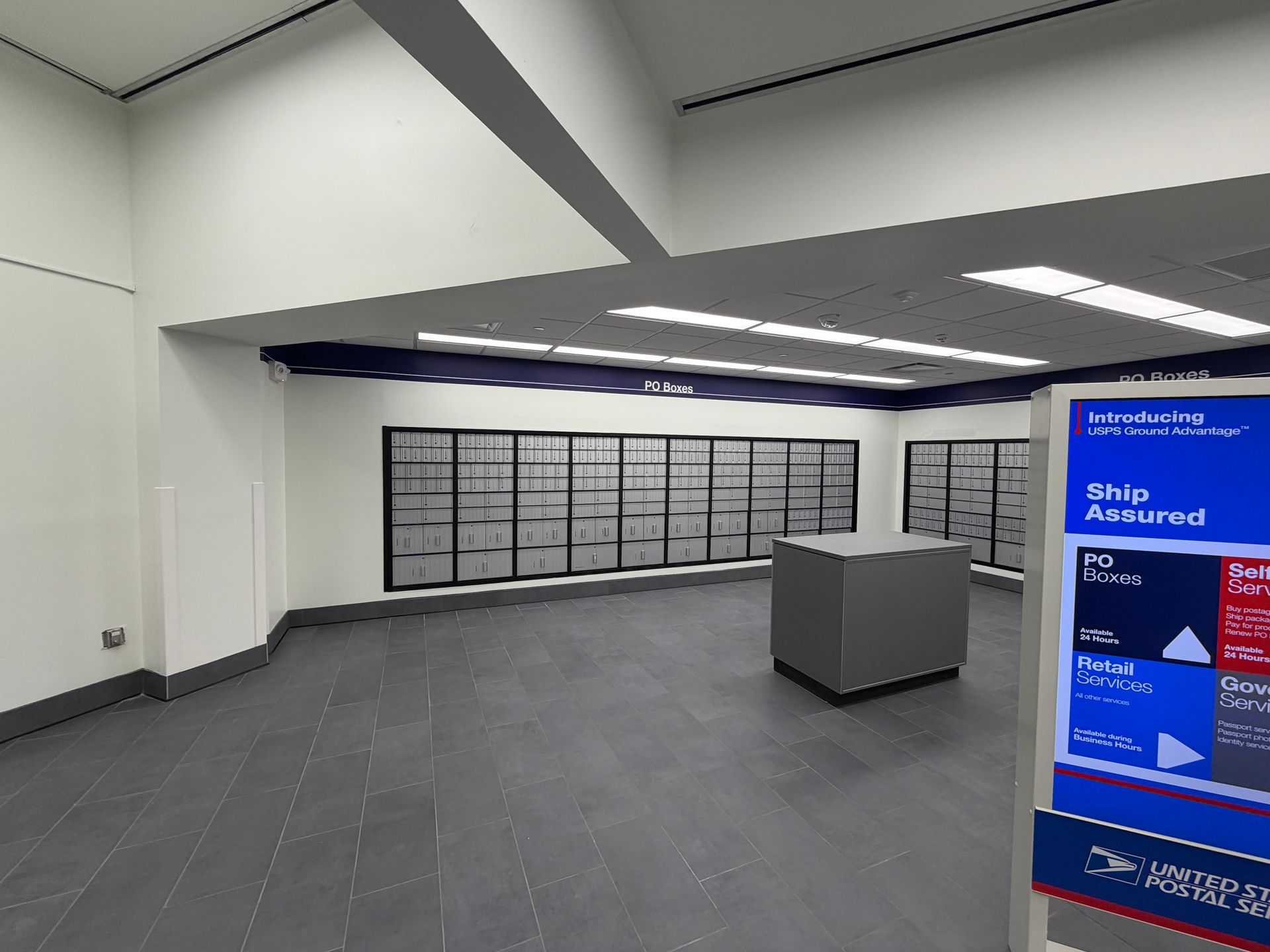 Interior of a USPS post office. Gray floor, rows of mailboxes, and a blue information display.