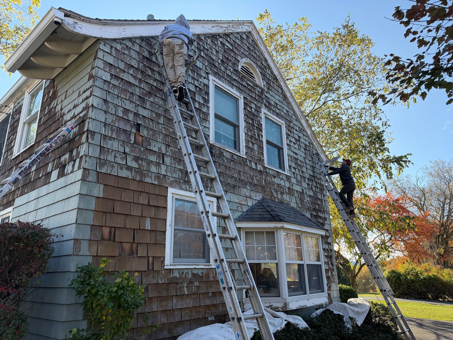 Two workers on ladders scraping peeling paint from the exterior of a two-story house.