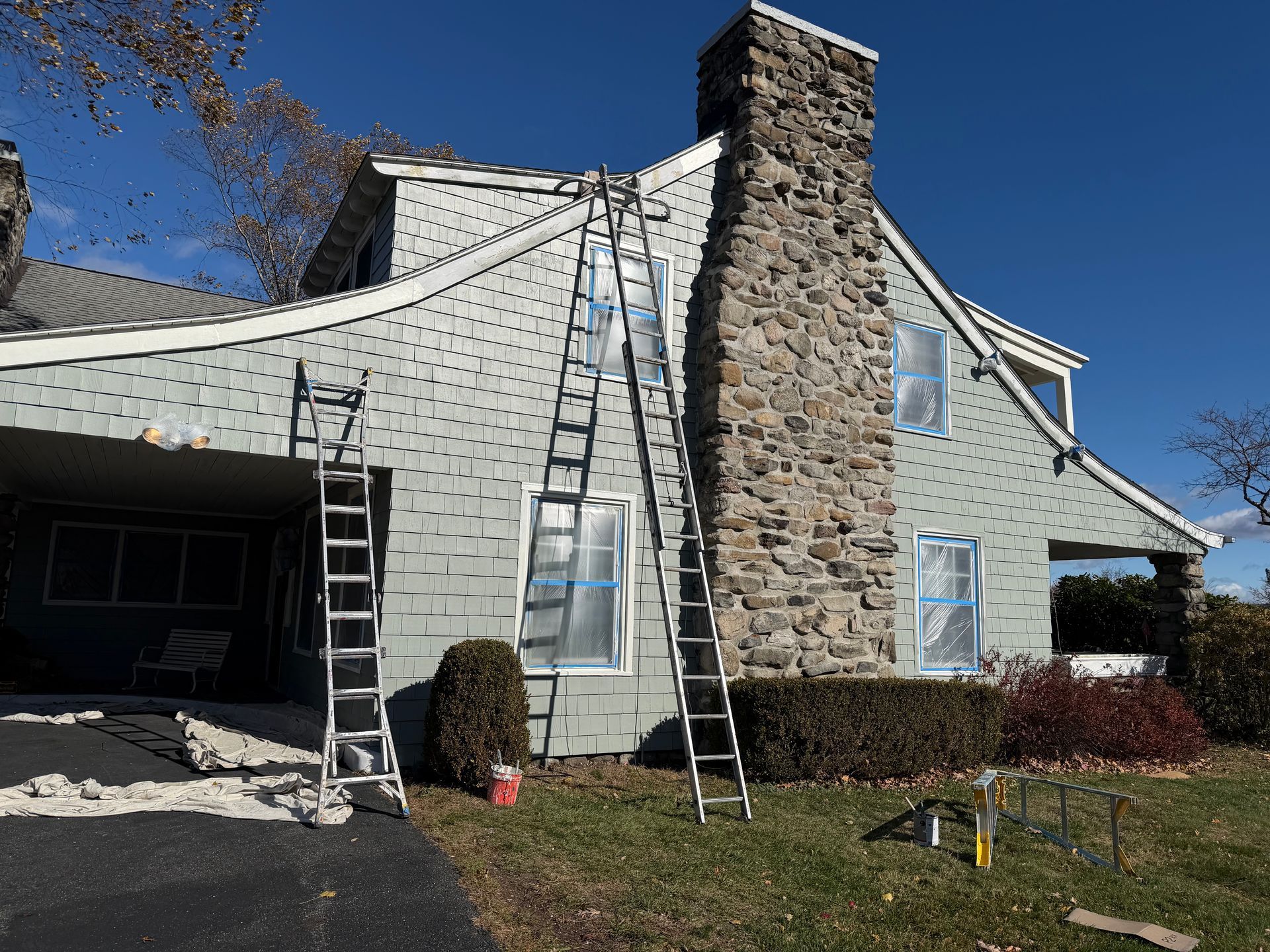 House exterior with ladders and chimney, windows taped for painting. Blue siding, stone chimney, clear sky.