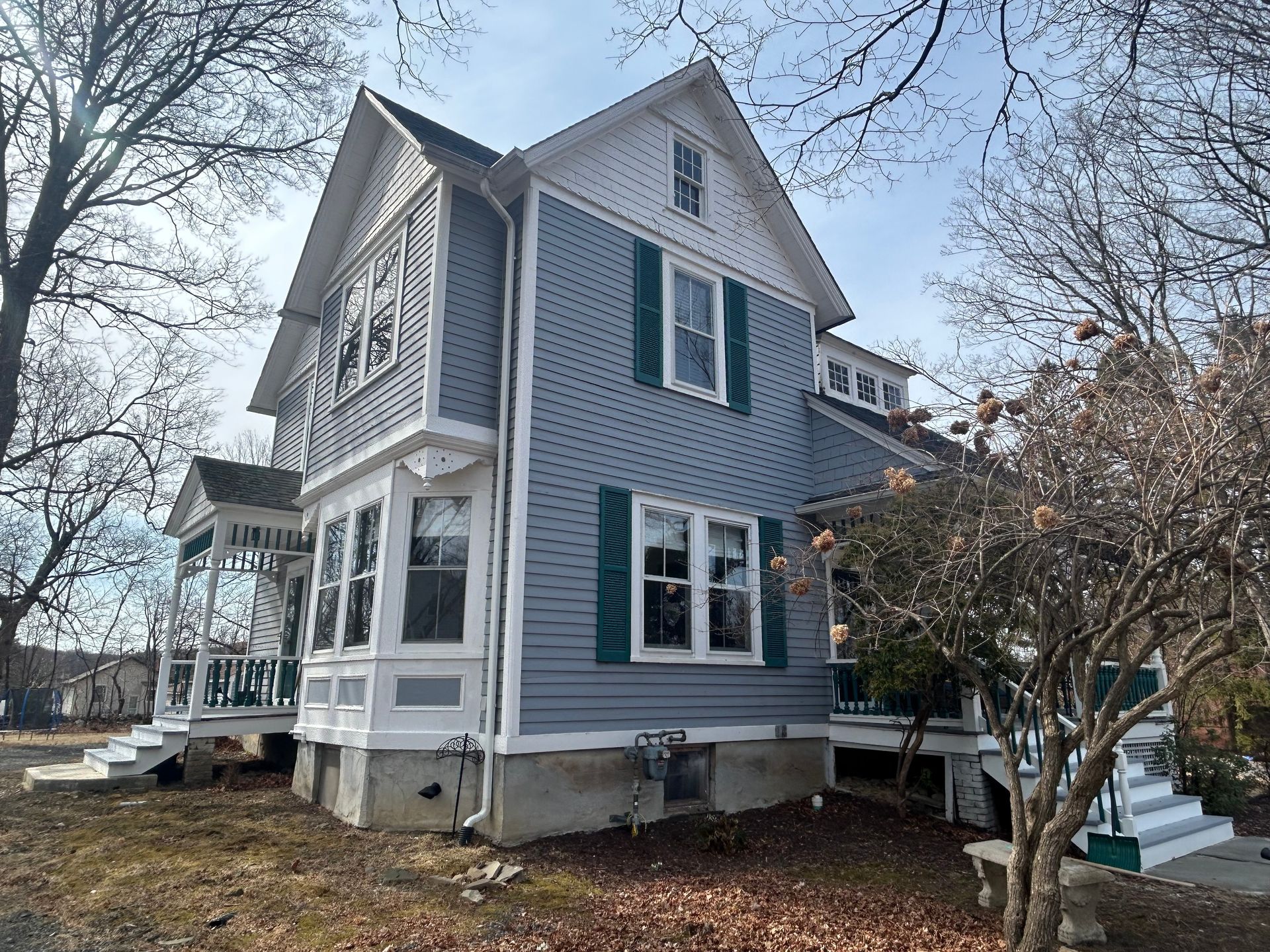 Blue house with white trim, green shutters, and a porch. Bare trees and blue sky.