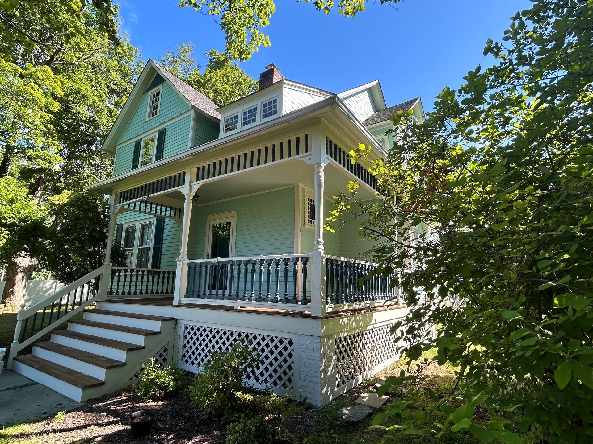 Two-story house with green siding, white trim, and a porch, surrounded by trees under a blue sky.