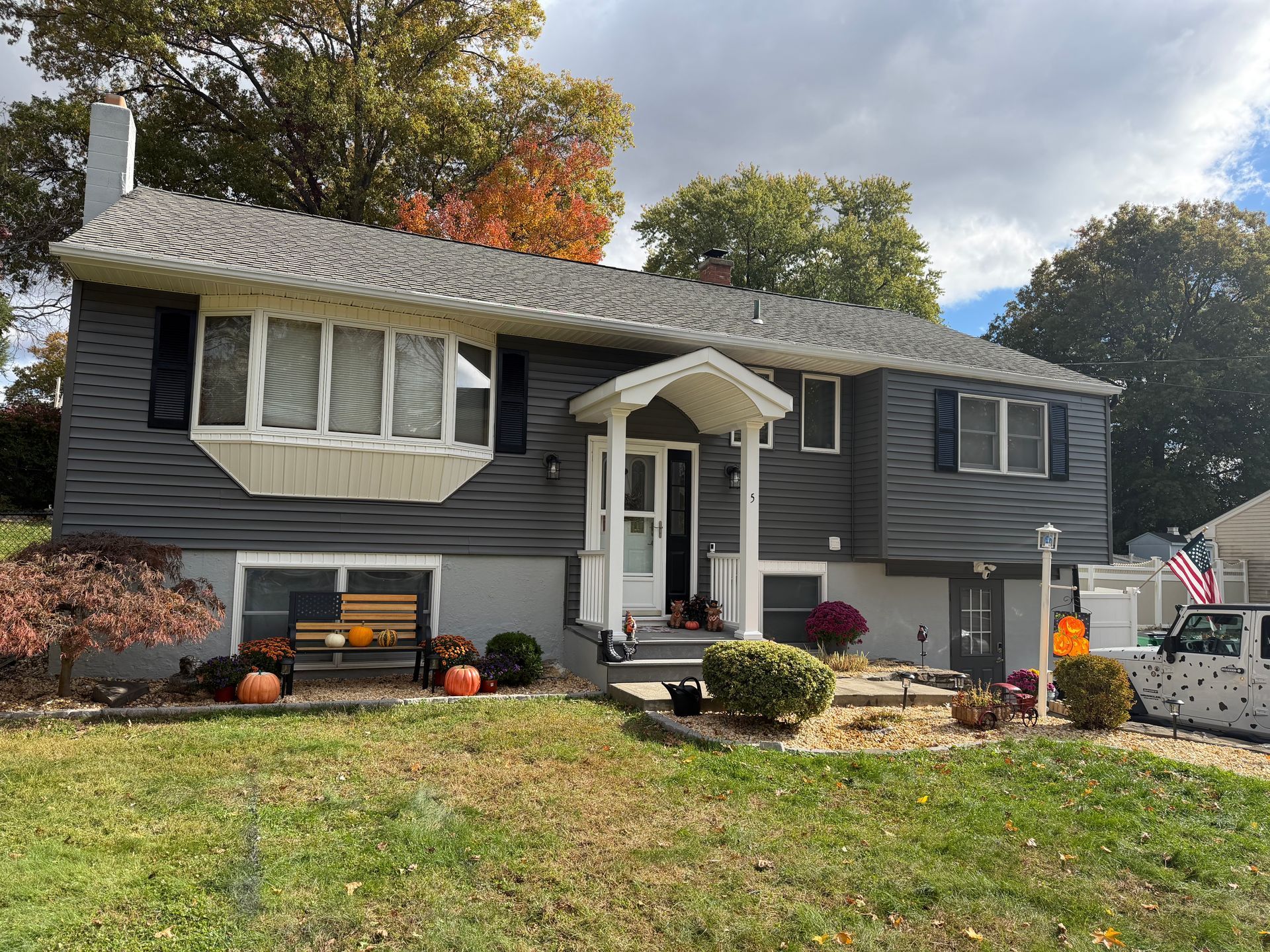 Gray house with fall decorations and an American flag on a lawn under a cloudy sky.