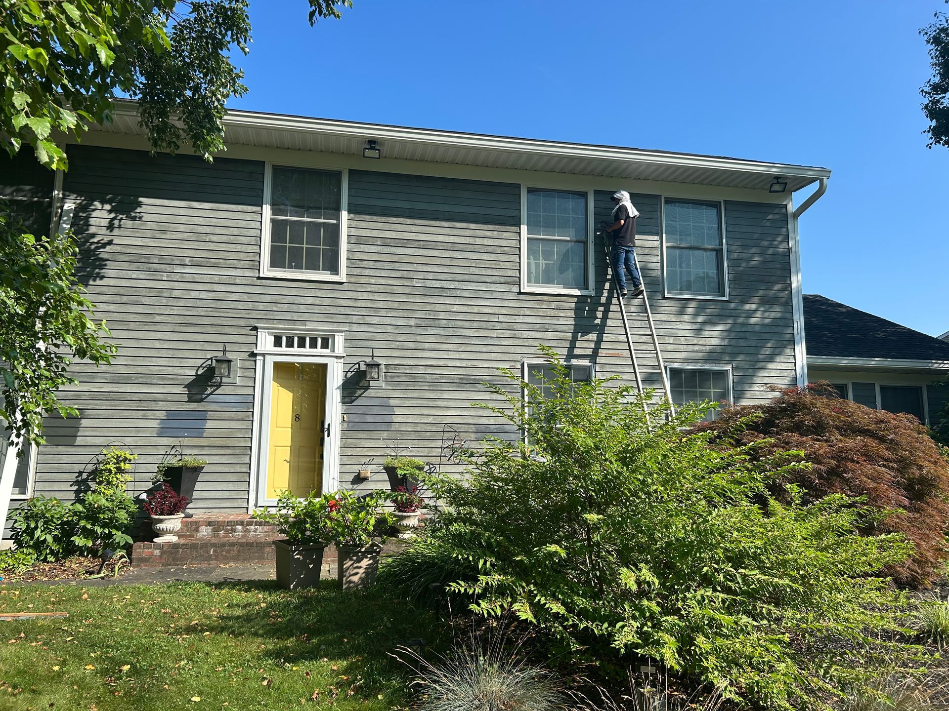 A person on a ladder is working on the siding of a two-story house with a yellow door, under a clear blue sky.