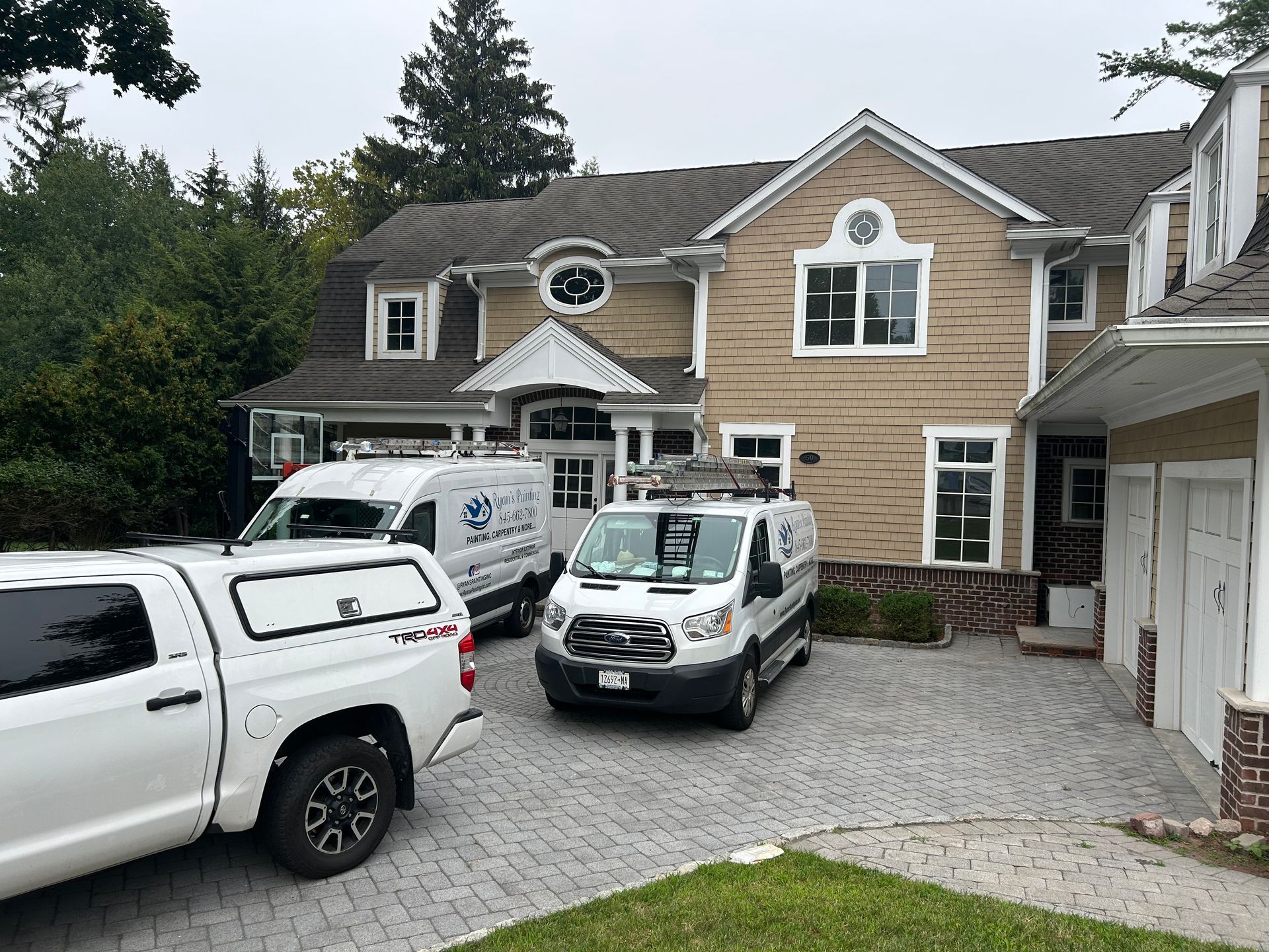 White trucks parked on a cobblestone driveway in front of a tan two-story house with a cloudy sky.