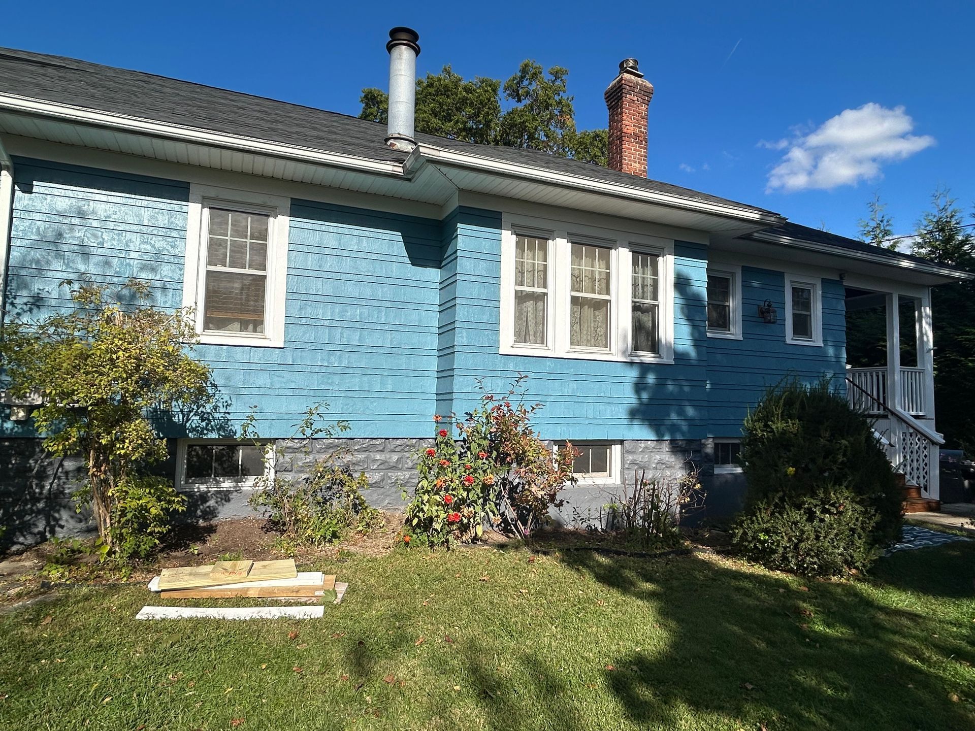 Blue house with white trim, windows, and chimney on a sunny day. Green grass and shrubs in foreground.