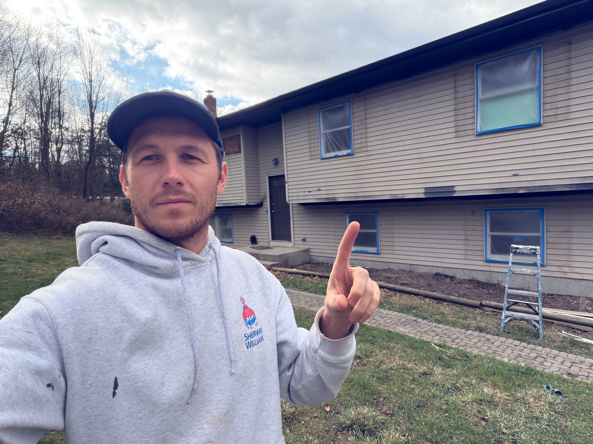 Man in gray hoodie points at a two-story house with blue-taped windows, cloudy sky.