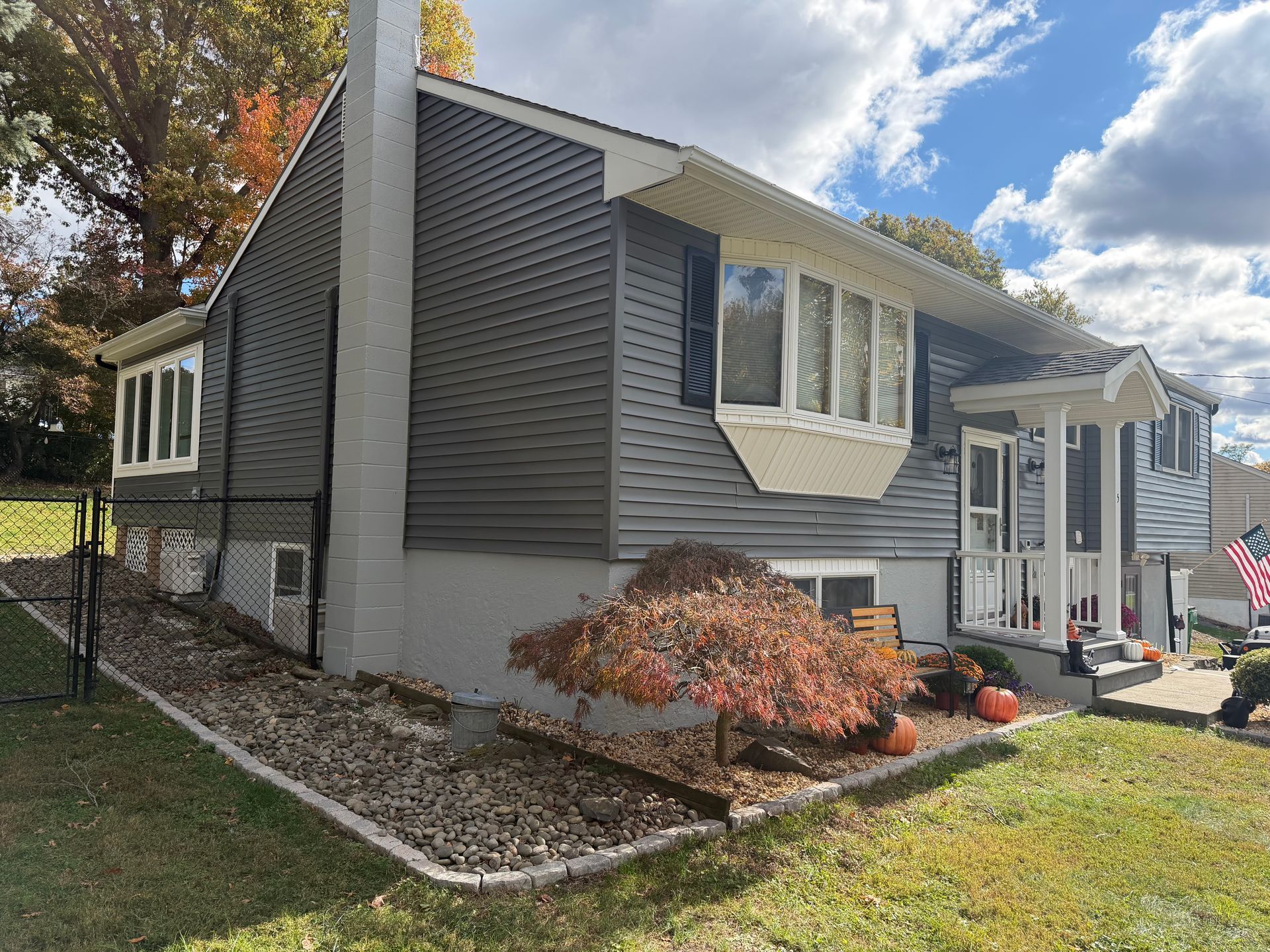 Gray house with a chimney and white trim, featuring a bay window, a small porch, and autumn foliage.