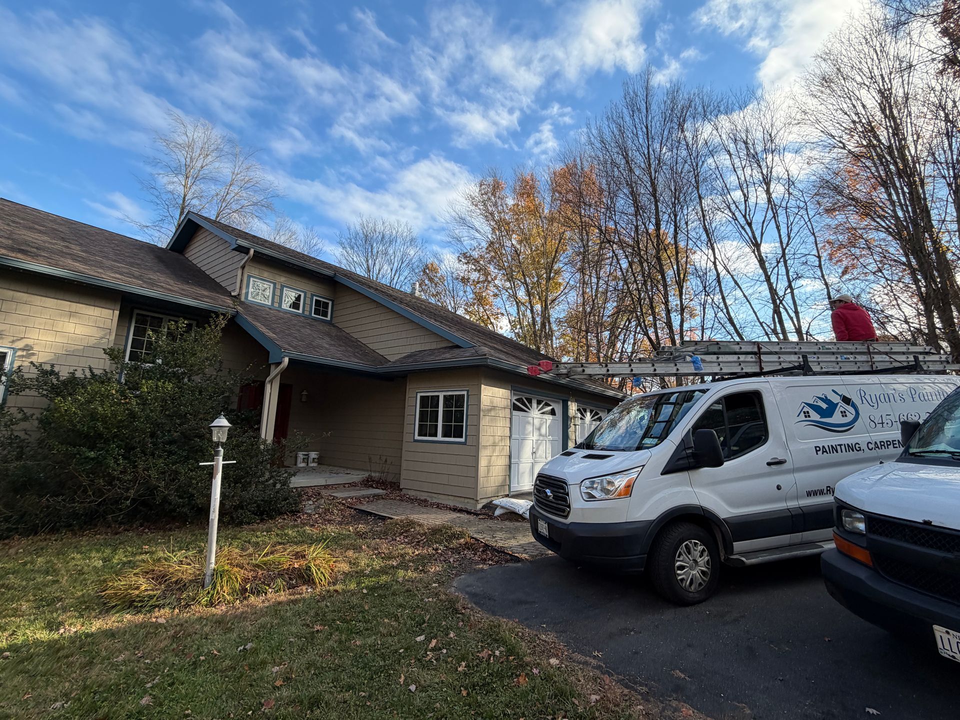 White van parked in front of a house. Two people, a ladder on roof, under a cloudy sky.