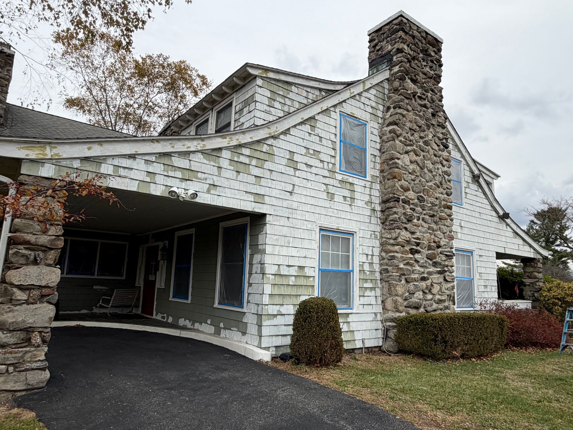 House with peeling white paint, stone chimney, and covered driveway. Asphalt driveway. Overcast sky.