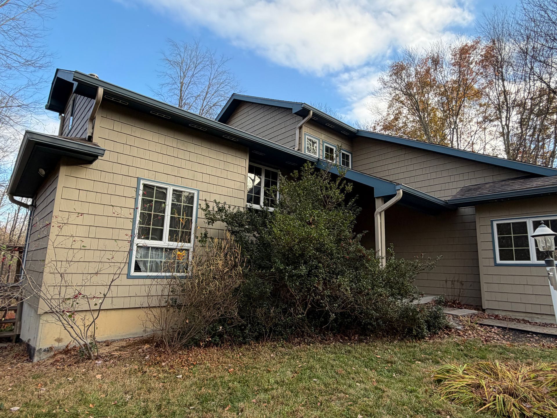 Beige house with blue roof and trim, several windows, and overgrown bushes in front.