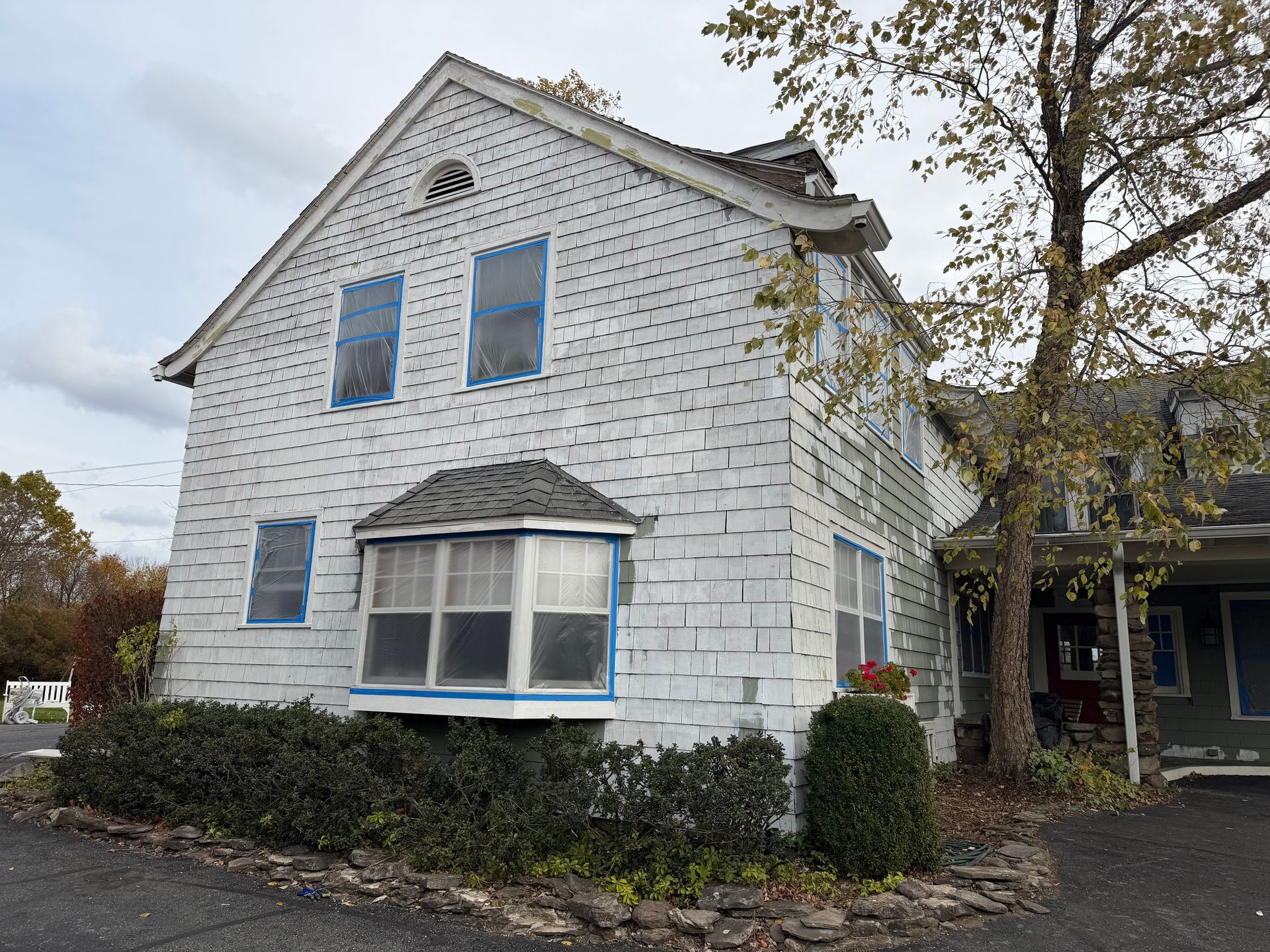 White two-story house with shingle siding, blue-taped windows, and a small tree beside it.