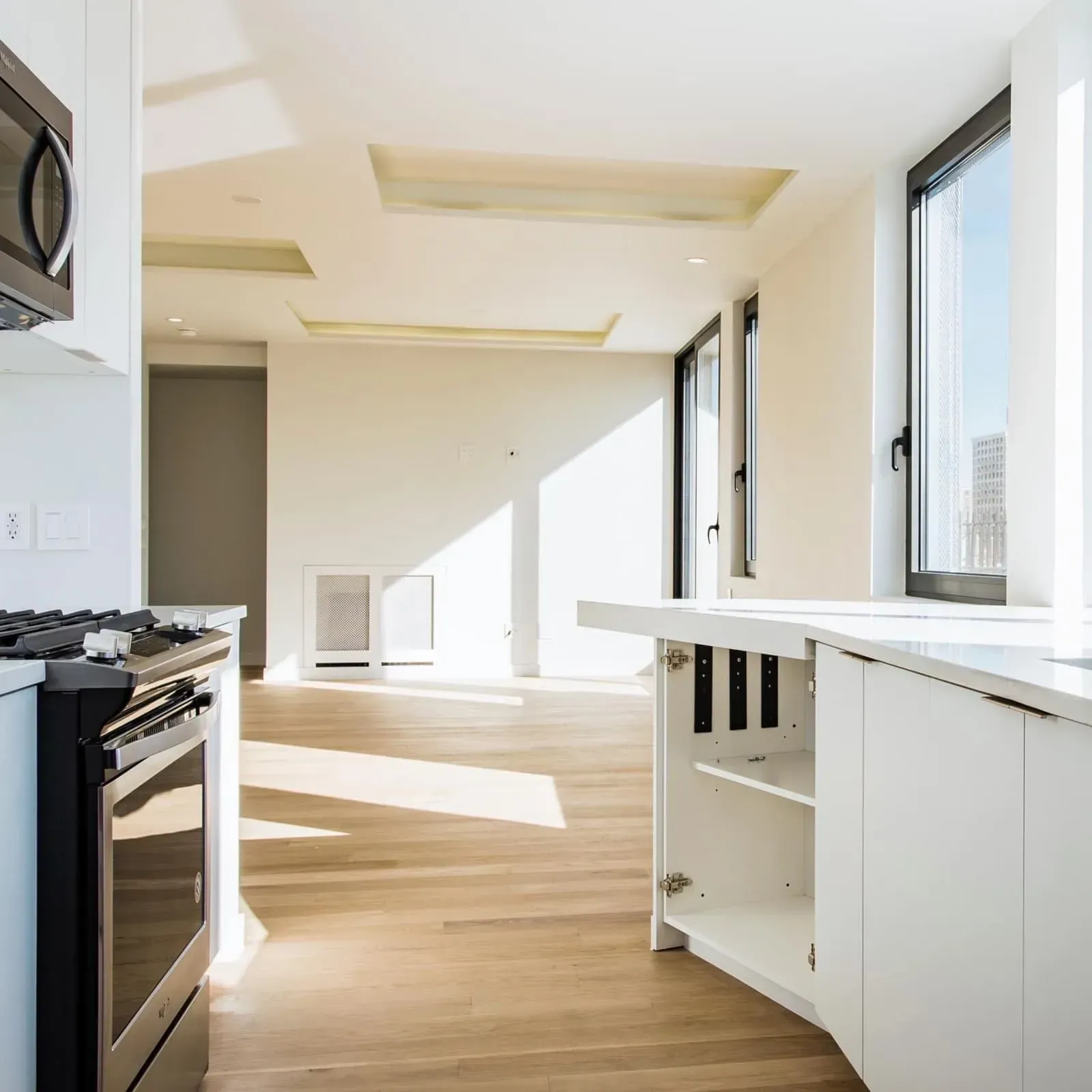 Bright white kitchen with light wood floors, oven, and counter. Sunlight streams through windows.
