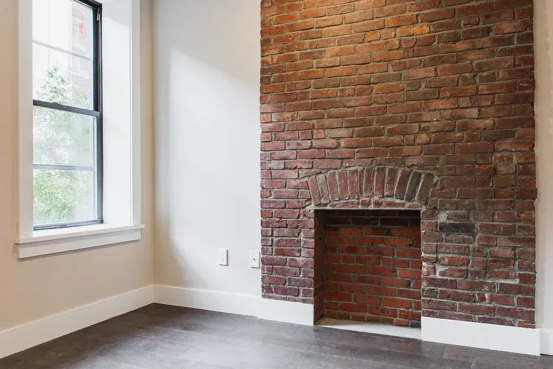 Empty room with dark hardwood floors, brick fireplace, and window.