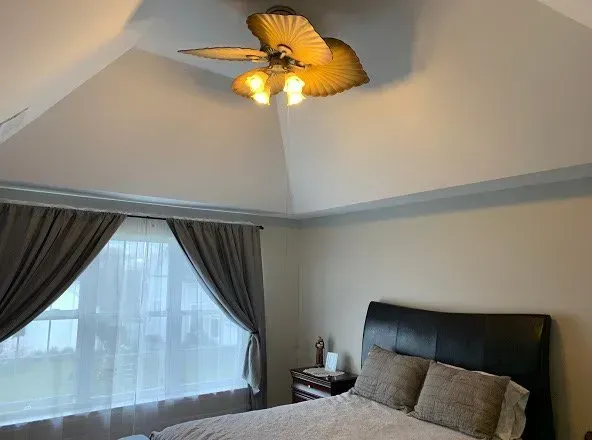 Bedroom with high ceiling, fan, bed, window with curtains. Soft lighting, neutral colors.