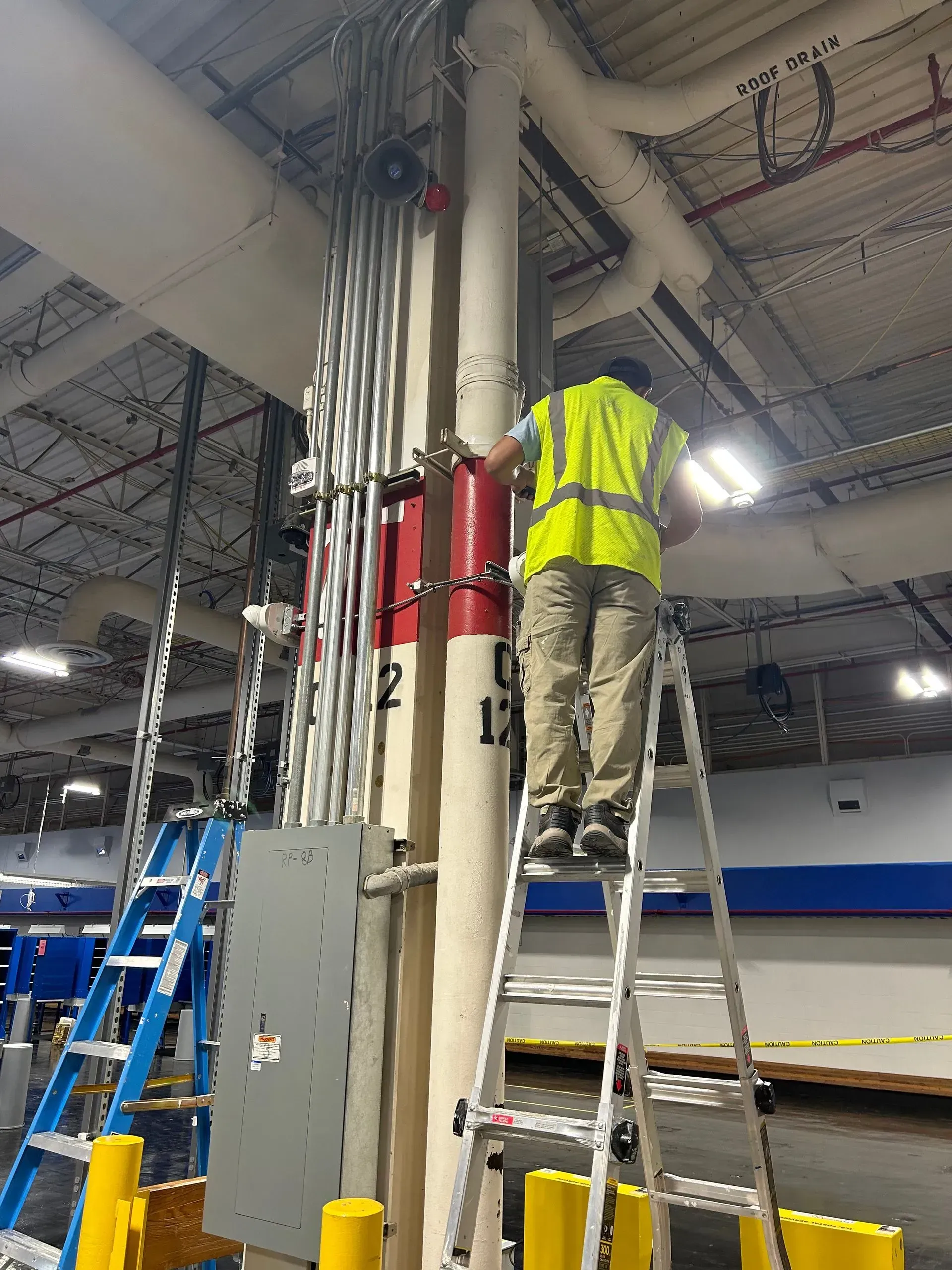 Worker in a safety vest on a ladder, installing electrical components on a support column inside a warehouse.