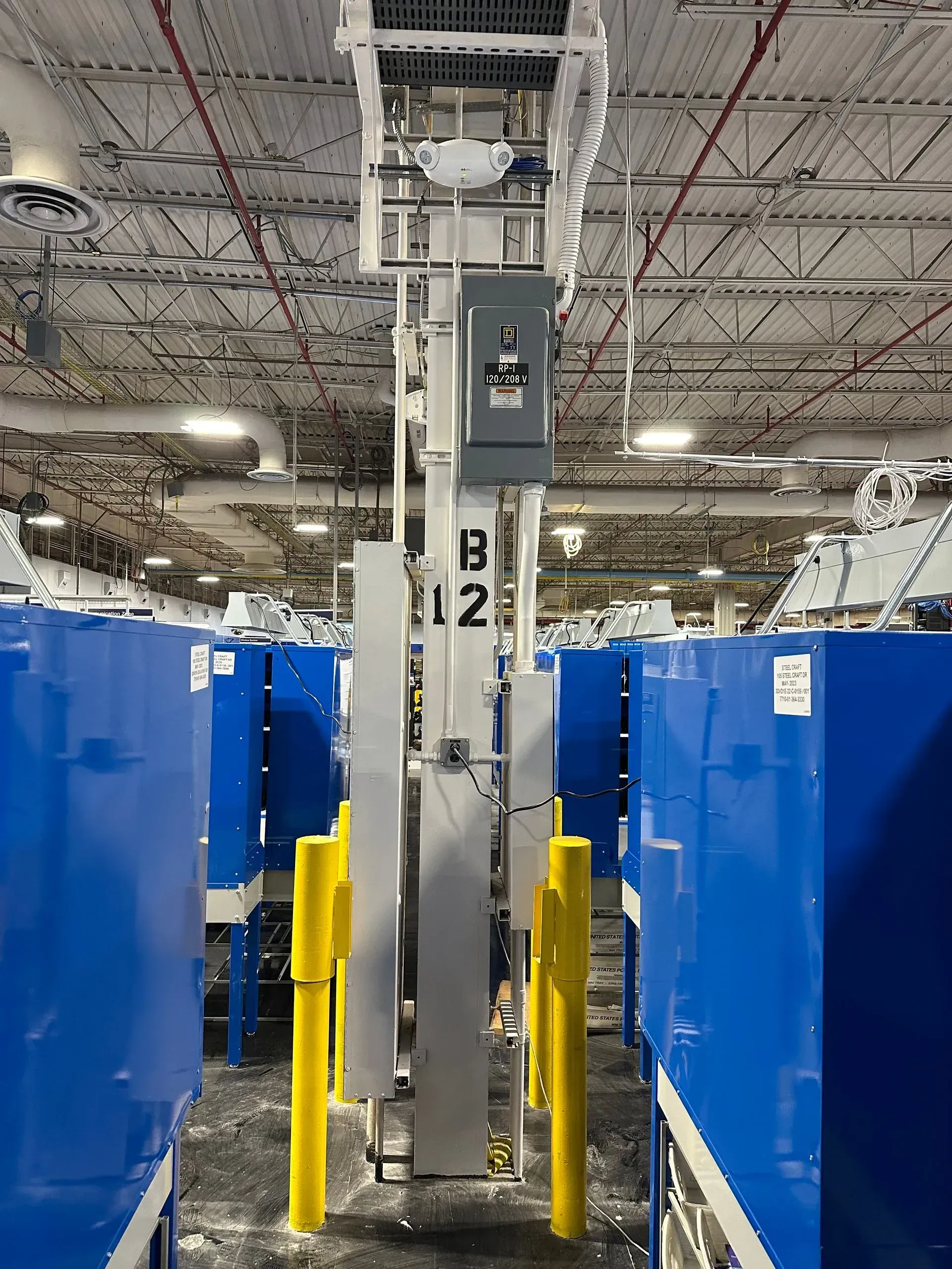 Warehouse scene with tall gray machinery between blue bins, yellow posts, and overhead lighting.