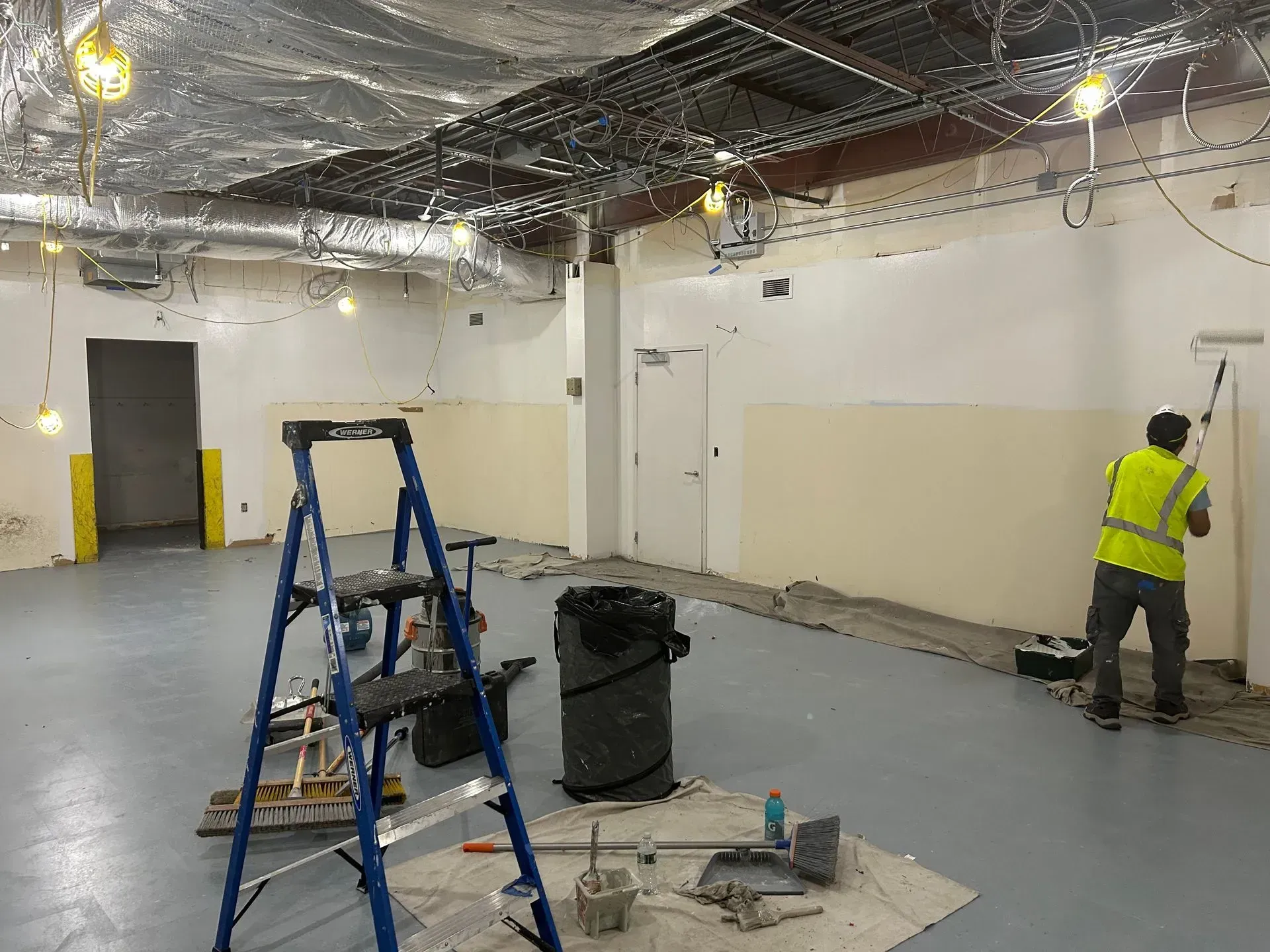 Man painting wall in a room under construction; includes a ladder, trash bin, and construction materials.