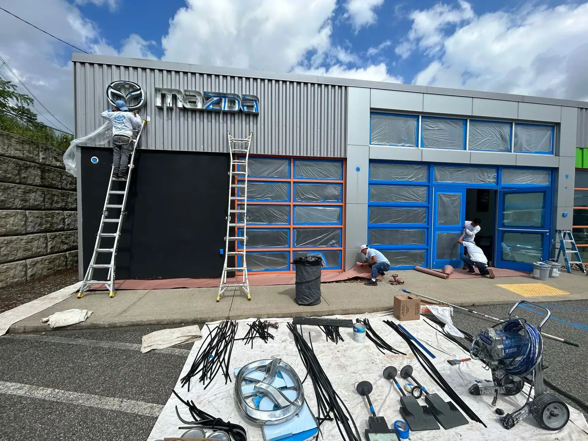 Workers painting a Mazda dealership building; blue accents and logo.