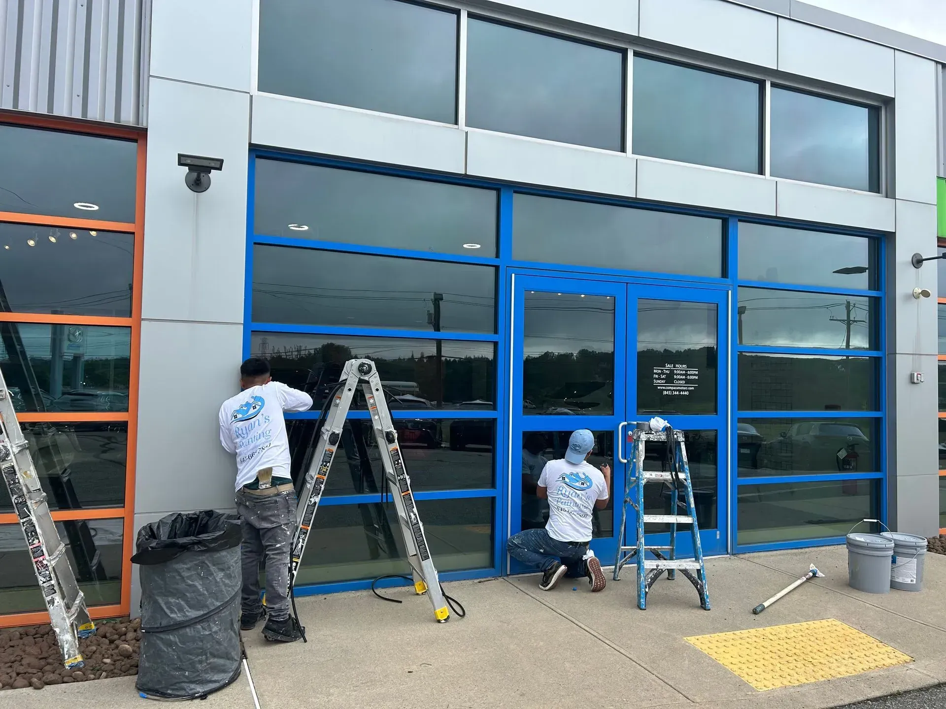 Two people installing window tint on a storefront with blue trim and large windows; ladders and supplies are present.