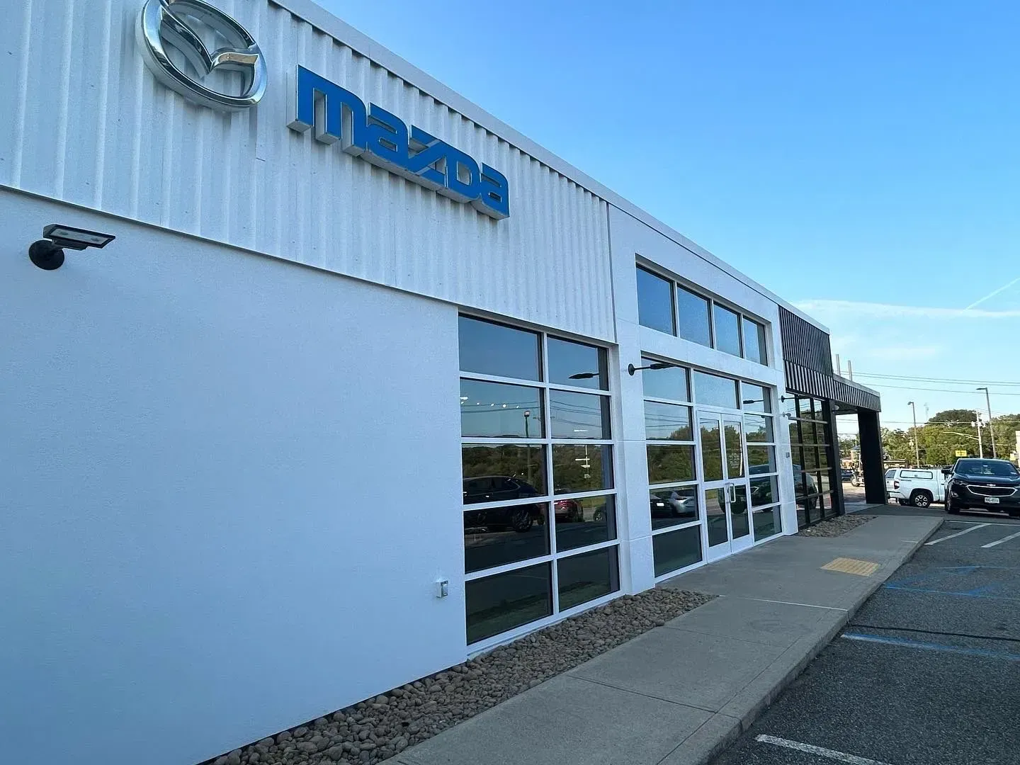Exterior view of a Mazda dealership with blue logo and large windows. The building is white and has a parking lot.