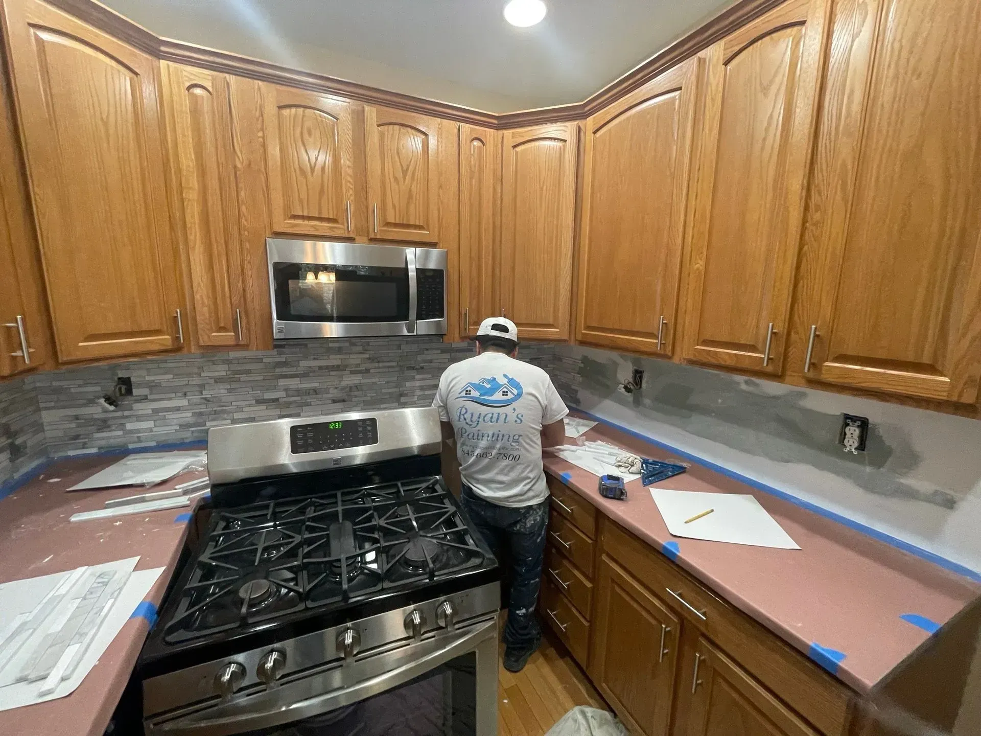 Person tiling a kitchen backsplash; brown cabinets, stainless steel appliances, pink countertop.