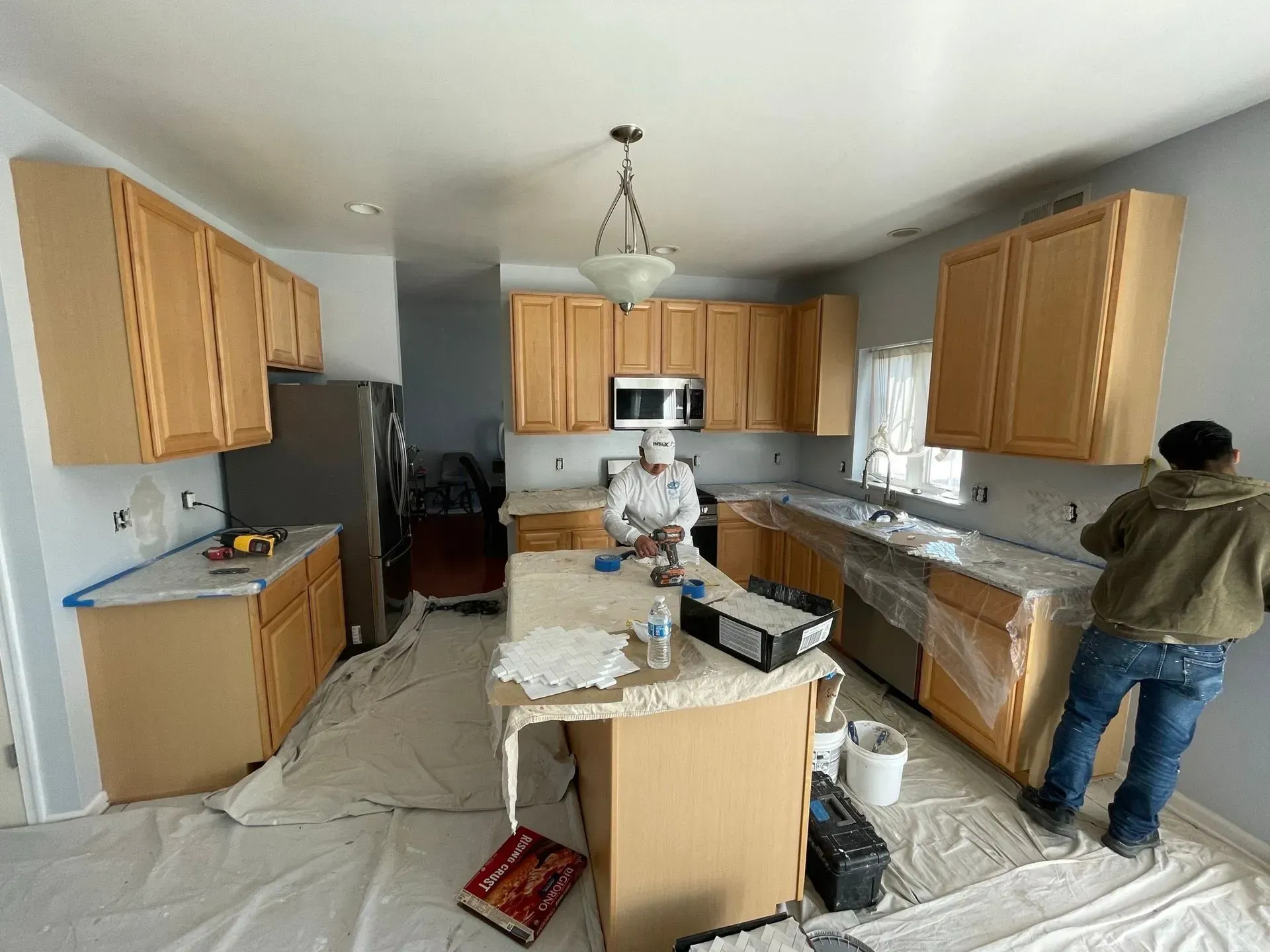 Two people painting a kitchen with light wood cabinets and countertops. Walls are being painted light blue.