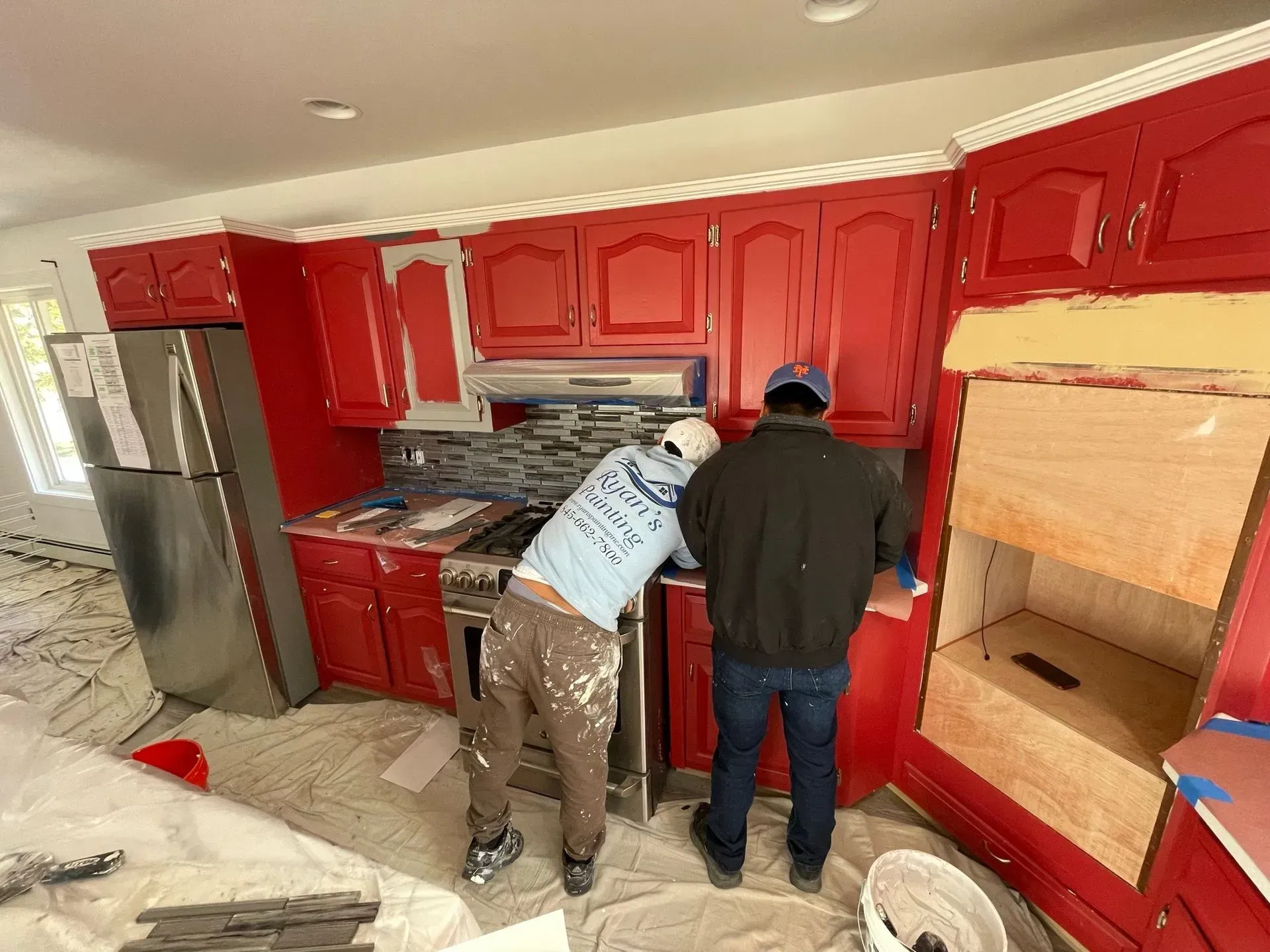 Two people painting red kitchen cabinets; one uses a brush.