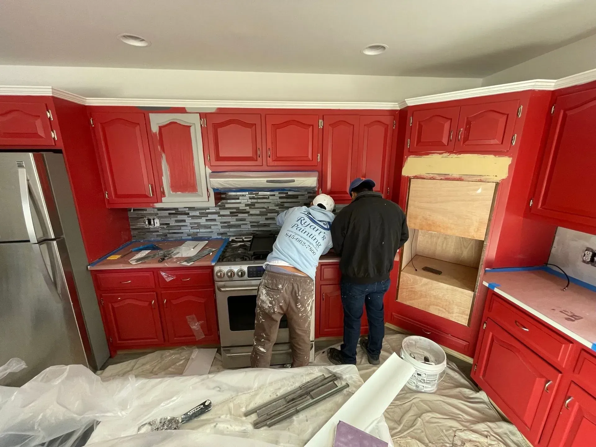 Two people painting red kitchen cabinets; one preps a cabinet above the stove.