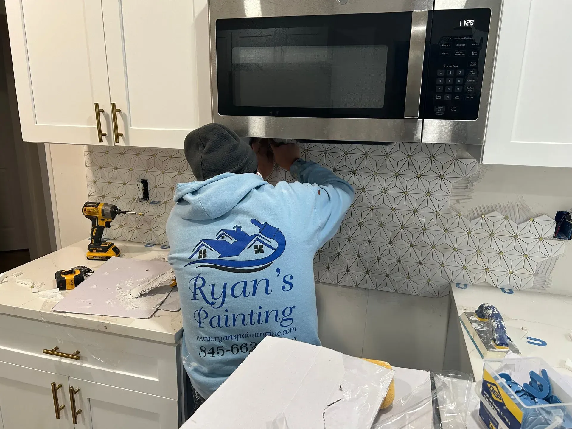 Person installing backsplash tile in a kitchen, beneath a microwave. He wears a light blue sweatshirt.