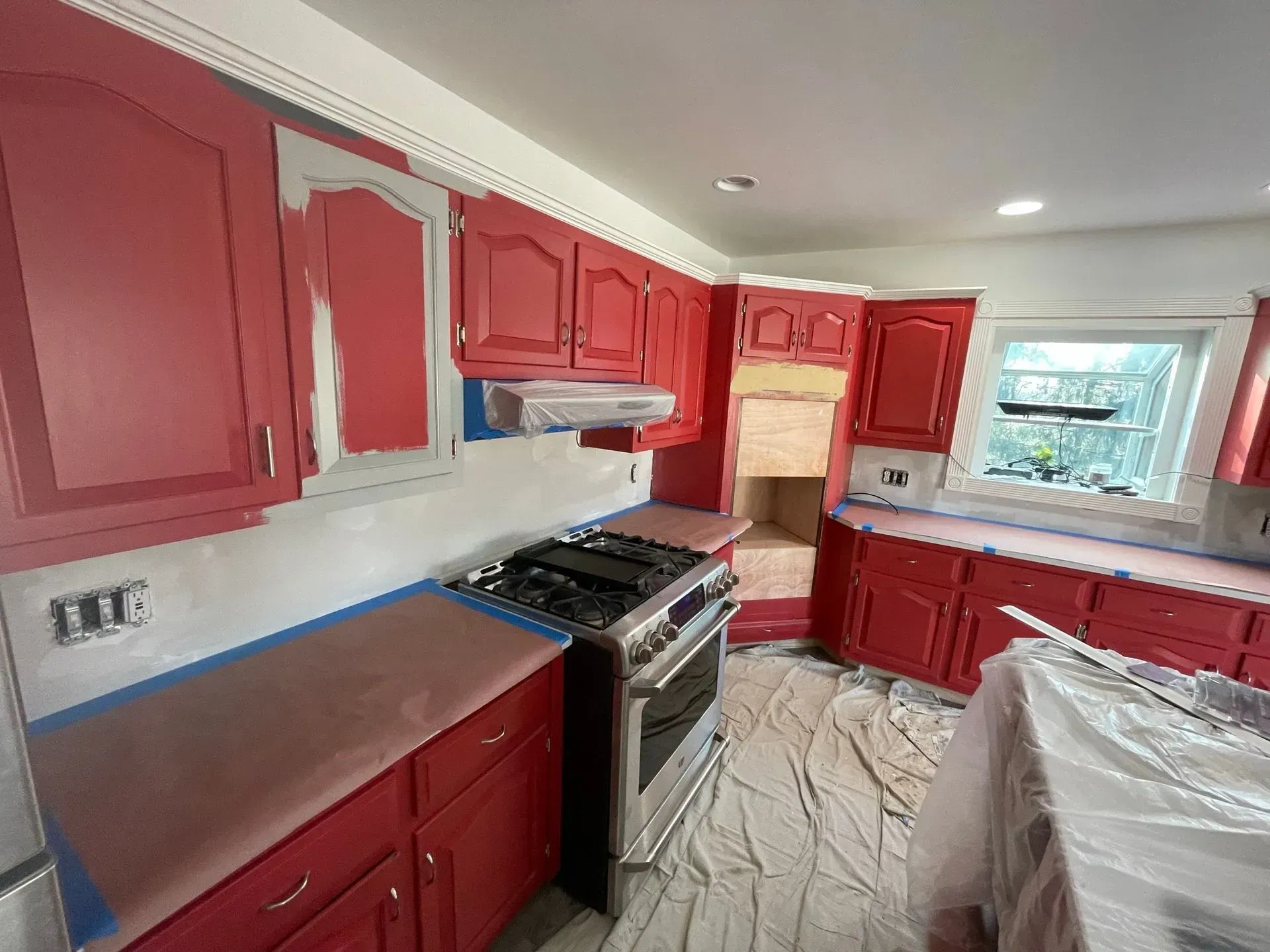 Kitchen with red cabinets being painted. Grey primer showing through, counters and appliances covered.