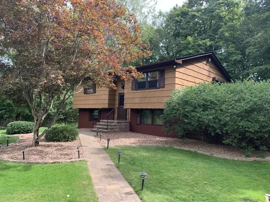 Two-story house with brown siding, pathway to front door, surrounded by greenery.