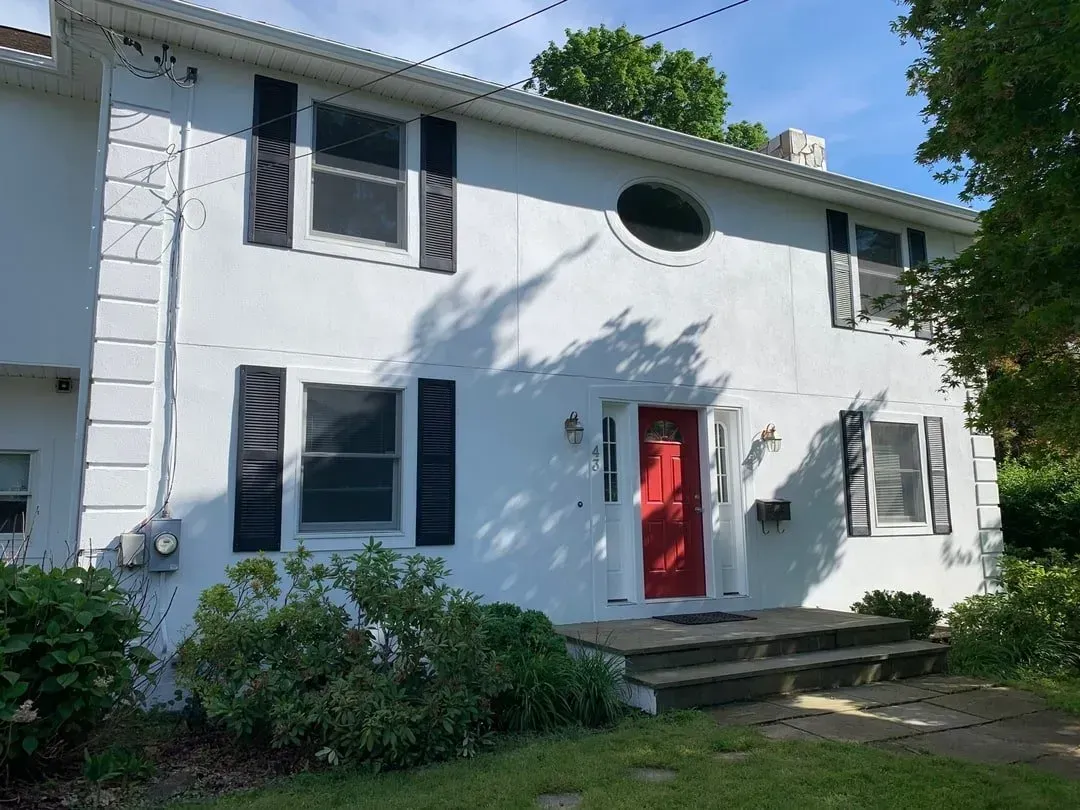 White two-story building with black shutters, red door, and small front yard with bushes.