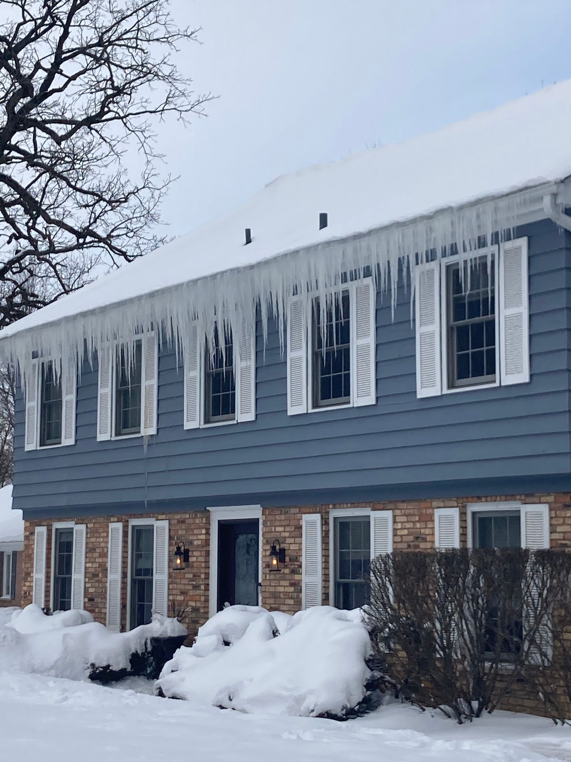 Two-story house with blue siding, white shutters, and large icicles hanging from the roof in a snowy setting.