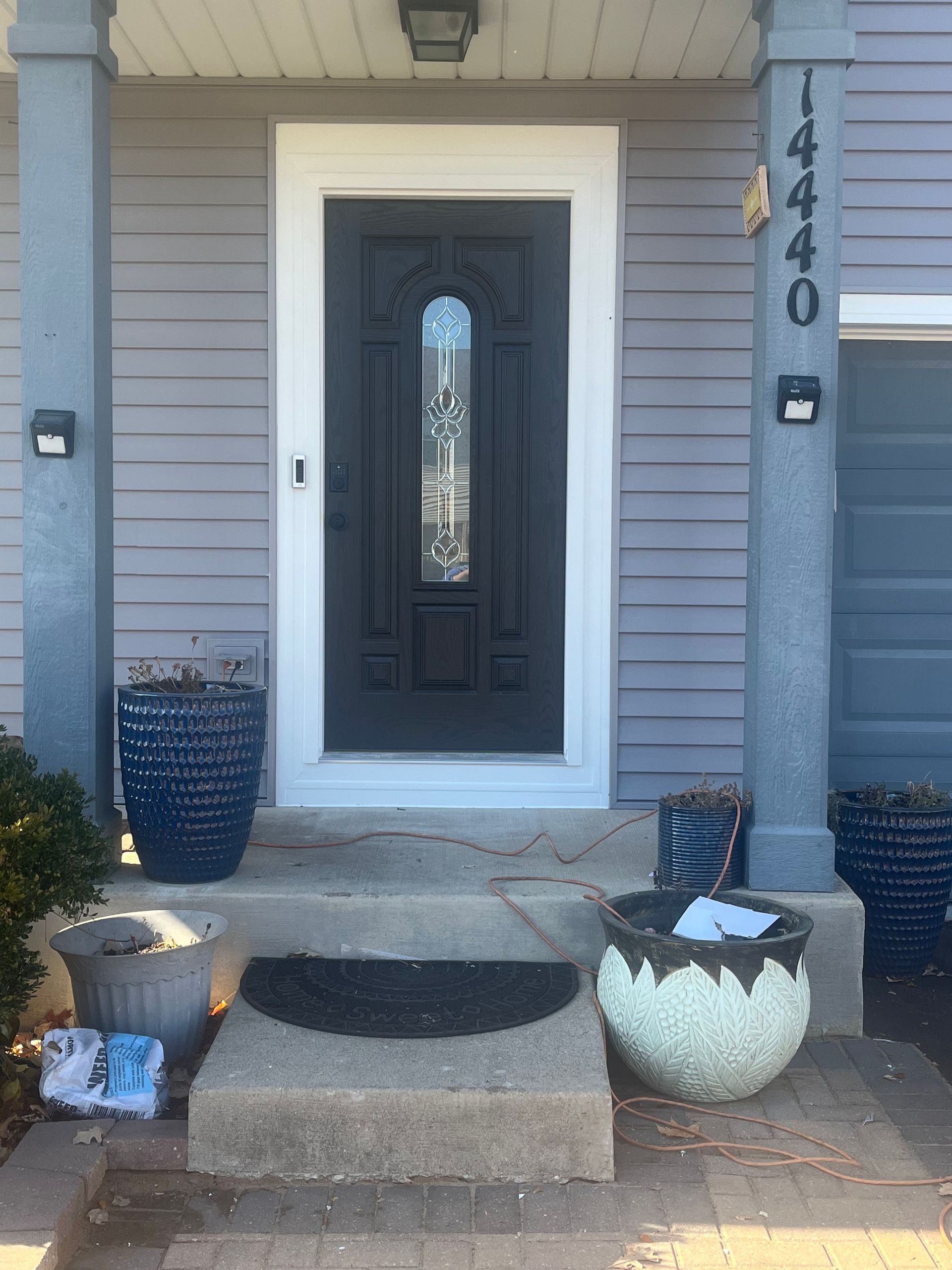 Gray house entrance with black door, white trim, and potted plants. Door has stained glass. Address numbers 
