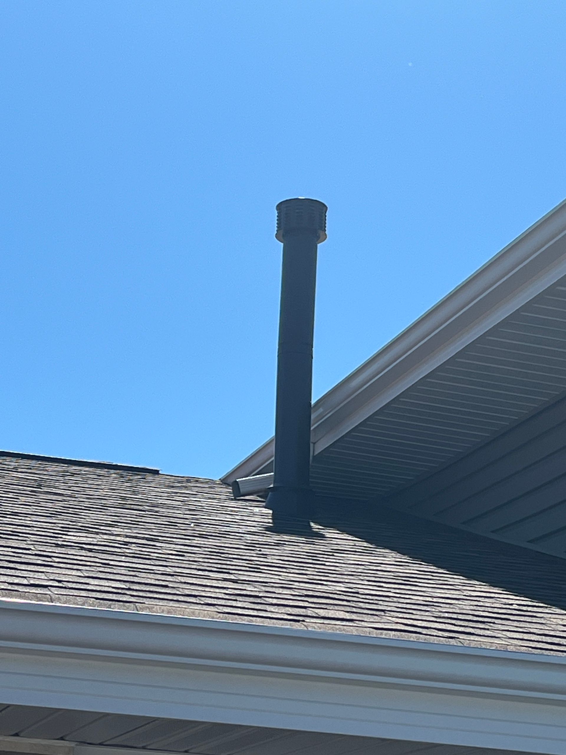 Black metal chimney protruding from a dark gray shingle roof against a blue sky.