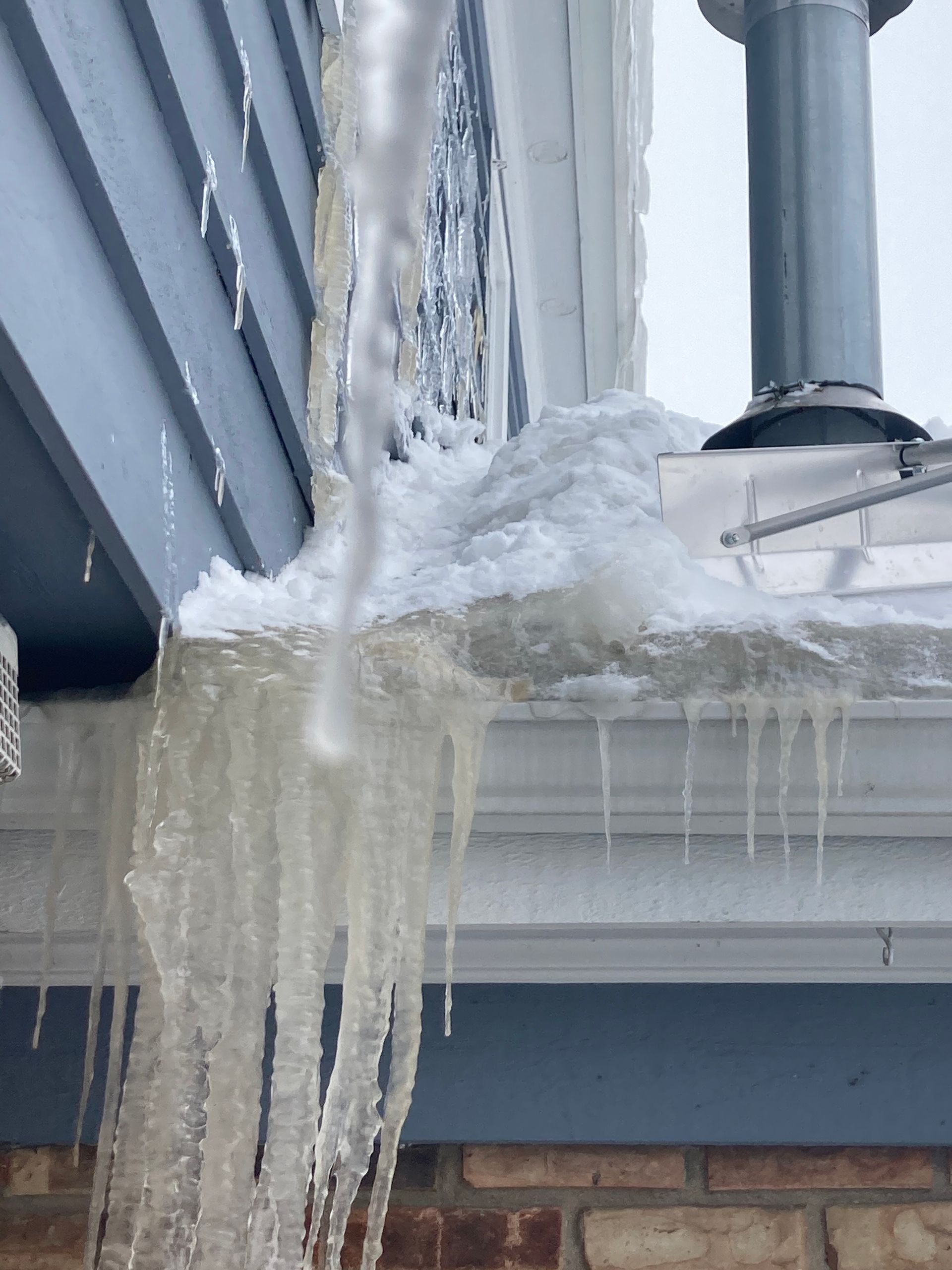 Icicles hanging from a roof with ice buildup near a chimney and blue siding.