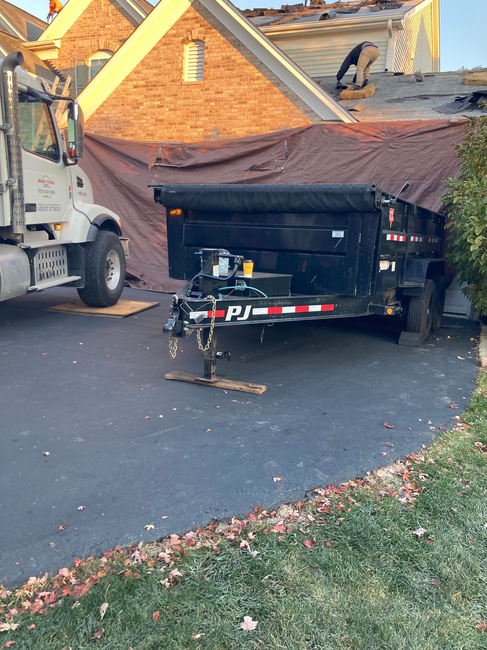 A dump truck and trailer beside a house under roof repair, covered by a tarp.